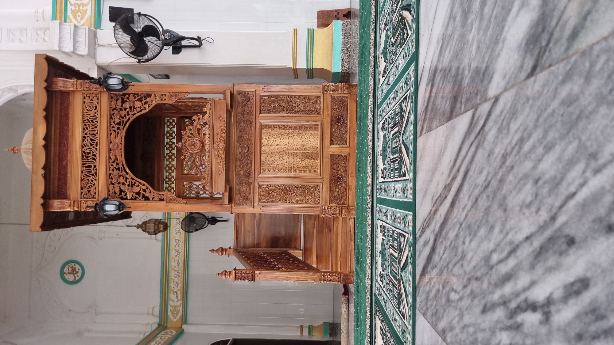 A low-angle, full shot captures a wooden minbar, likely in a mosque in Lapang Timu, Indonesia. The intricately carved minbar, made of light-colored wood, stands prominently in the center of the frame. It features decorative paneling, arches, and finials, with a tiered roof topped by a dome. A wooden staircase leads up to the platform. Two black lanterns hang from the minbar, and two black electric fans are mounted on the white wall behind it. A green prayer rug with a repeating pattern is laid out on the floor in front of the minbar. The floor itself is made of polished marble with a grey and white veined pattern. The background reveals white walls with decorative trim in green and gold. A circular emblem with Arabic script is visible on the upper left wall. The overall scene is one of a clean and well-maintained religious space.