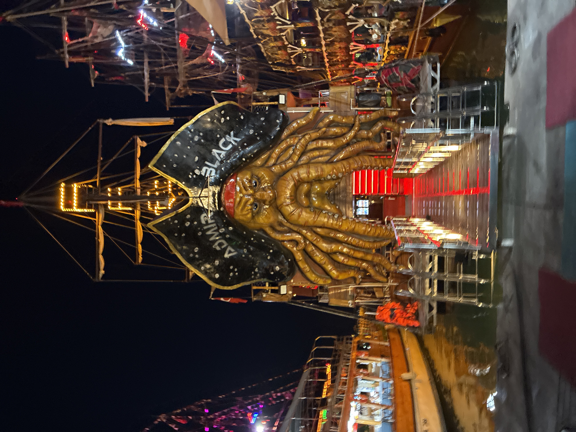 This image shows a nighttime scene at what appears to be an amusement park or carnival in Alanya, Turkey. The main subject is a large, elaborate float or attraction designed to resemble a pirate ship. Dominating the center is a prominent Kraken-like figure with a human-like face, tentacles, and wearing a red bandana. The figure is a golden-brown color and appears to be made of a sculpted material. Behind the Kraken, a large black flag, possibly part of the ship's sail, is visible with white speckles and the words "BLACK" and "ADMIRAL" printed on it.
The attraction is brightly lit with numerous yellow and red lights, illuminating the intricate rigging and mast of the pirate ship. Metal scaffolding and platforms are visible around the attraction, suggesting it's a complex structure, possibly for a ride or performance. The reflection of the lights can be seen on a wet surface below, likely water, indicating the attraction might be situated near a harbor or on a platform over water.
In the background, other carnival elements are suggested by colorful lights and structures, hinting at a lively atmosphere. The overall mood is festive and visually exciting, characteristic of a nighttime entertainment venue. The image is oriented vertically, with the primary focus on the pirate ship attraction.