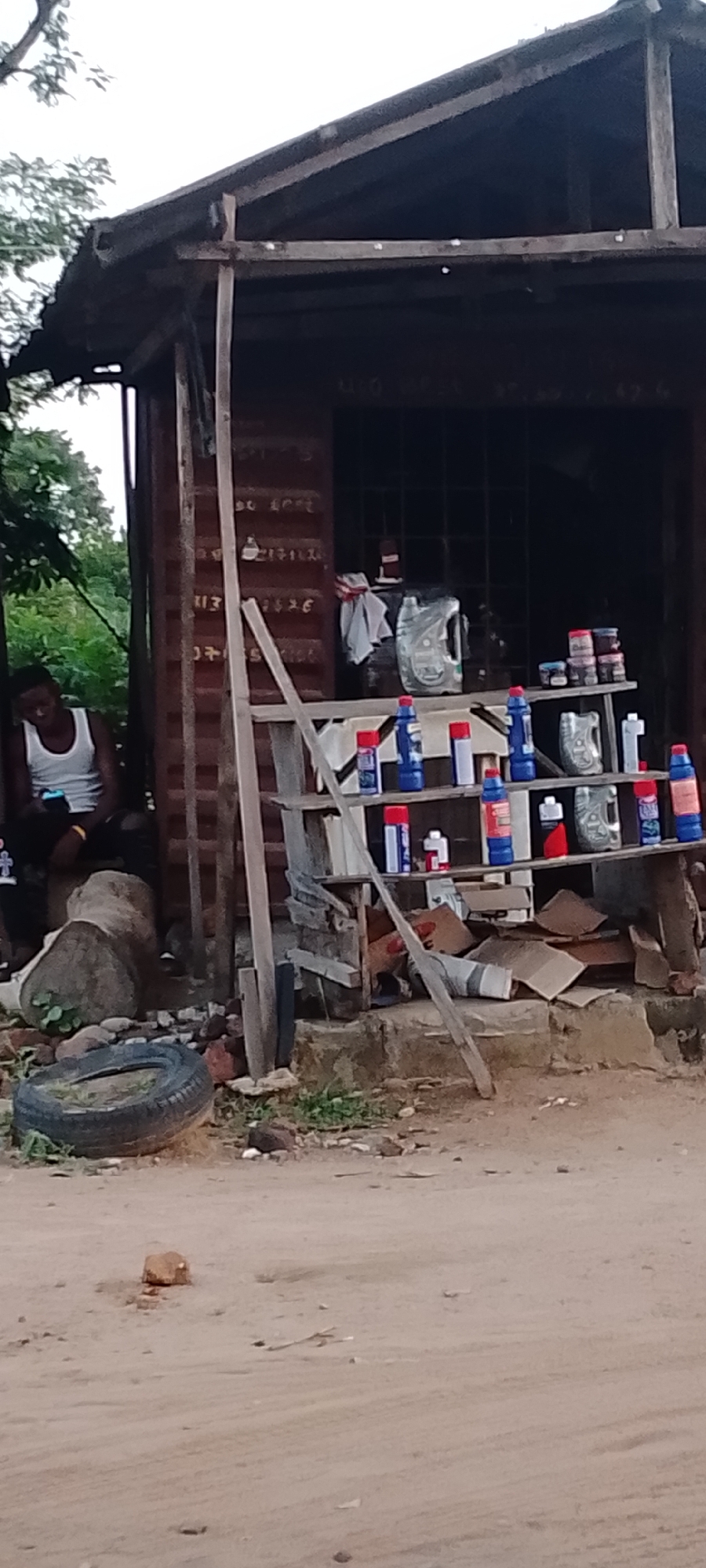 The image depicts a makeshift roadside shop in Adankolo, Nigeria. A man in a white tank top and dark pants sits on a log, holding a mobile phone. Behind him is a corrugated metal structure, likely a shipping container repurposed as a shop. A wooden two-tiered shelf displays various car care products, including oil containers and spray cans. The shop appears to be in a semi-rural area. A used tire lies on the ground near the log. The ground is dusty and sandy.