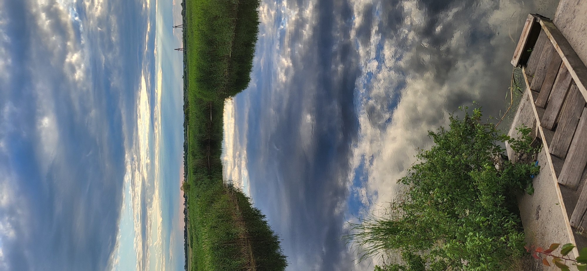 The image depicts a serene, natural landscape with a body of water mirroring the sky. The scene is dominated by the reflections of clouds on the water's surface, creating a dramatic contrast between dark and light gray, with hints of blue sky peeking through. Along the water's edge, dense green reeds or tall grass grow, forming a verdant border.
On the right side of the image, a wooden structure, possibly part of a dock or pier, is visible. It's made of weathered wooden planks and is positioned on a concrete bank. A lush green bush with dense foliage grows near this structure. The overall impression is one of natural beauty and tranquility.
The time of day appears to be late afternoon or early evening, suggested by the soft lighting and the mixture of light and dark clouds, which could indicate an approaching storm or the end of a sunny day. The weather seems to be partly cloudy, with dramatic cloud formations dominating the sky.
There is no visible text in the image. The location context of Hetmanivka, Ukraine, suggests a rural or semi-rural setting, consistent with the presence of water, reeds, and a natural landscape. The scene evokes a sense of peace and immersion in nature.