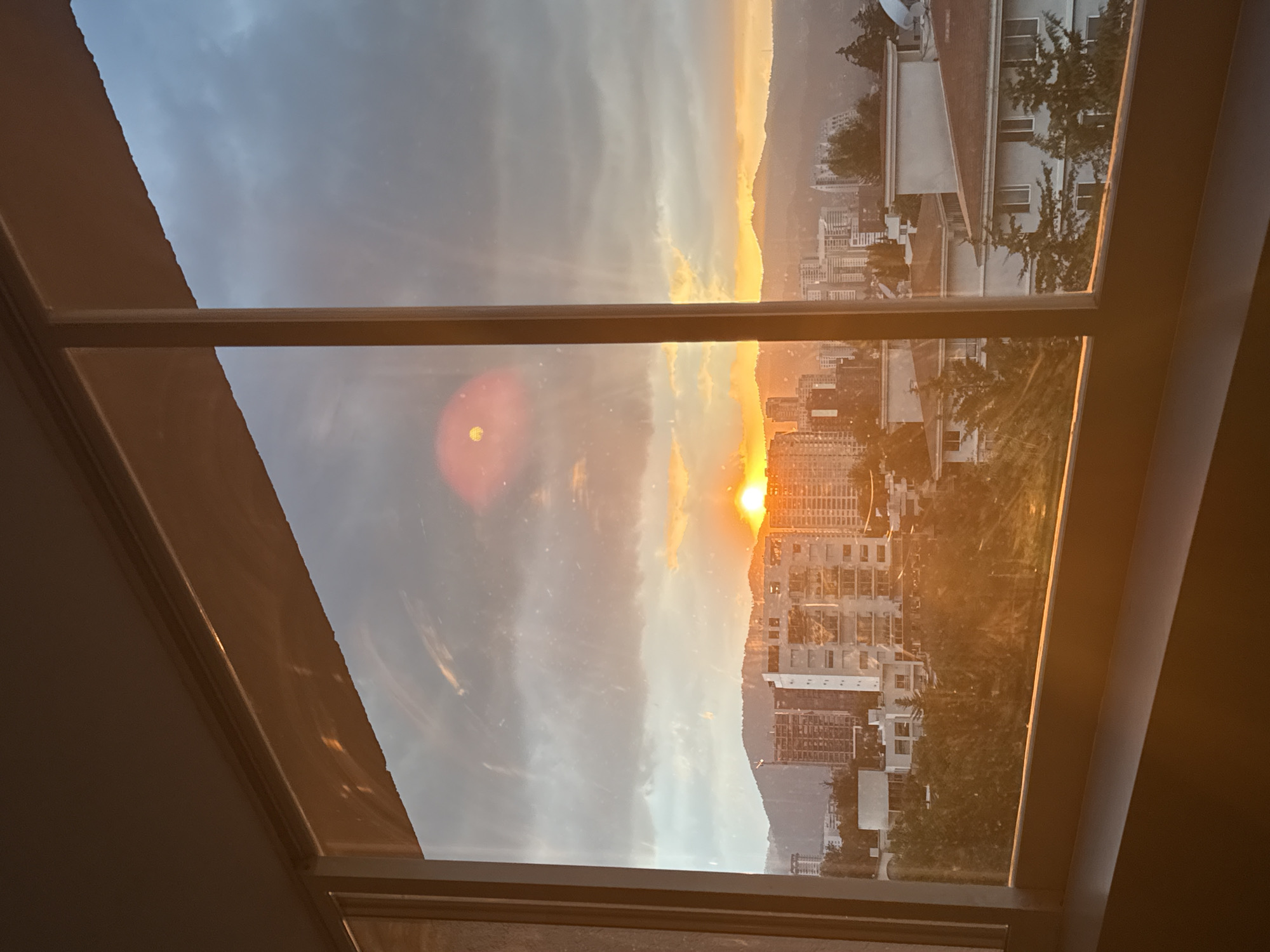 The image depicts a cityscape view through a window at sunset or sunrise. The sky is a mix of orange, yellow, and gray, with a bright sun visible between buildings. There is an urban landscape with buildings of varying heights, possibly located in Mexico City, Mexico. The window frame is visible, creating a border around the scene.
