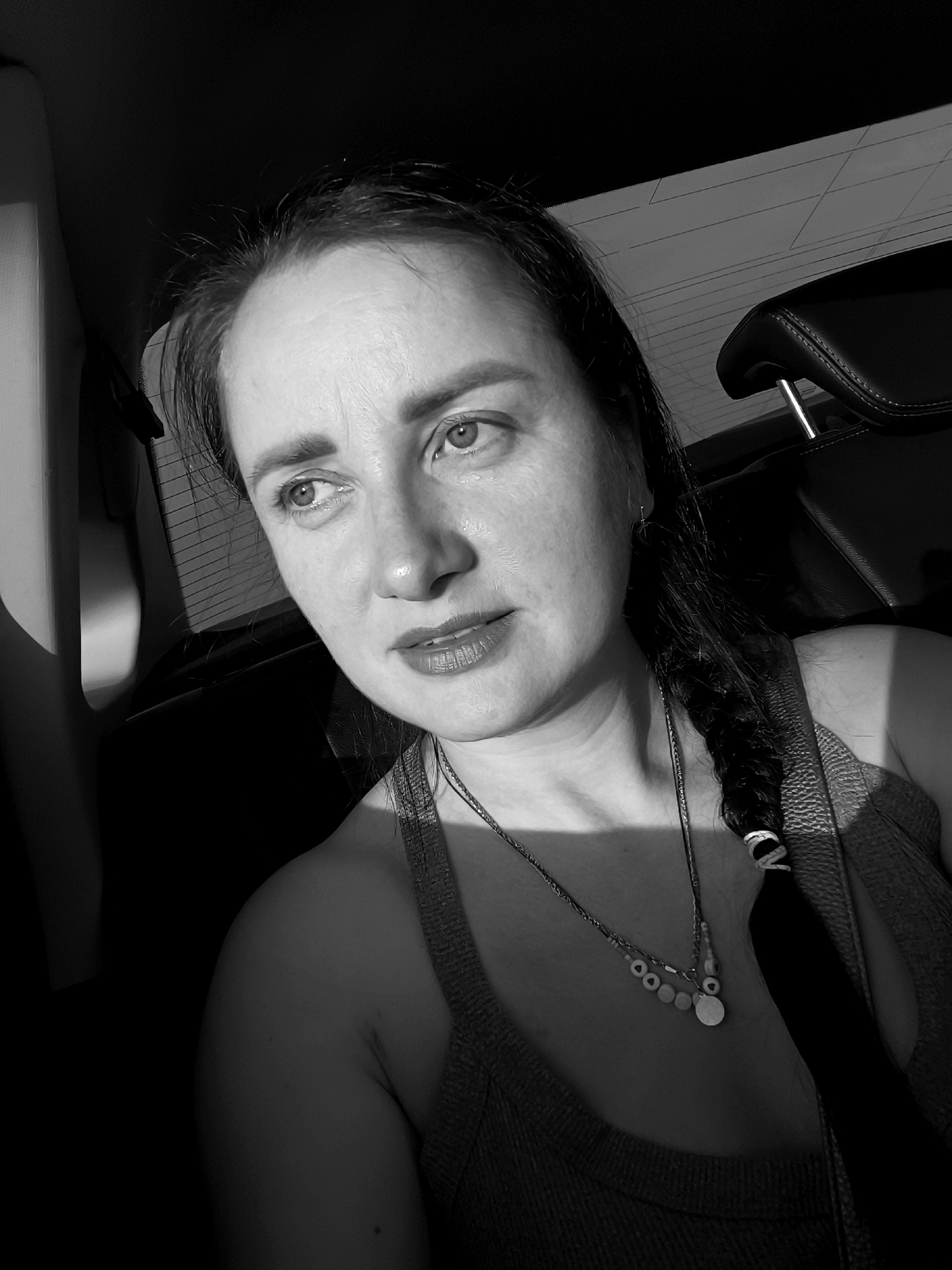 A black and white, close-up, selfie-style shot shows a woman with dark hair braided down her right side, sitting in the passenger seat of a vehicle. She is looking to her left, with her head slightly tilted. She is wearing a necklace with several beads and a pendant. The interior of the car, including a headrest and part of the back seat, is visible. The lighting suggests it is daytime, possibly late afternoon, with sunlight casting shadows on her face and the car interior. The image is from Lelystad, Netherlands.