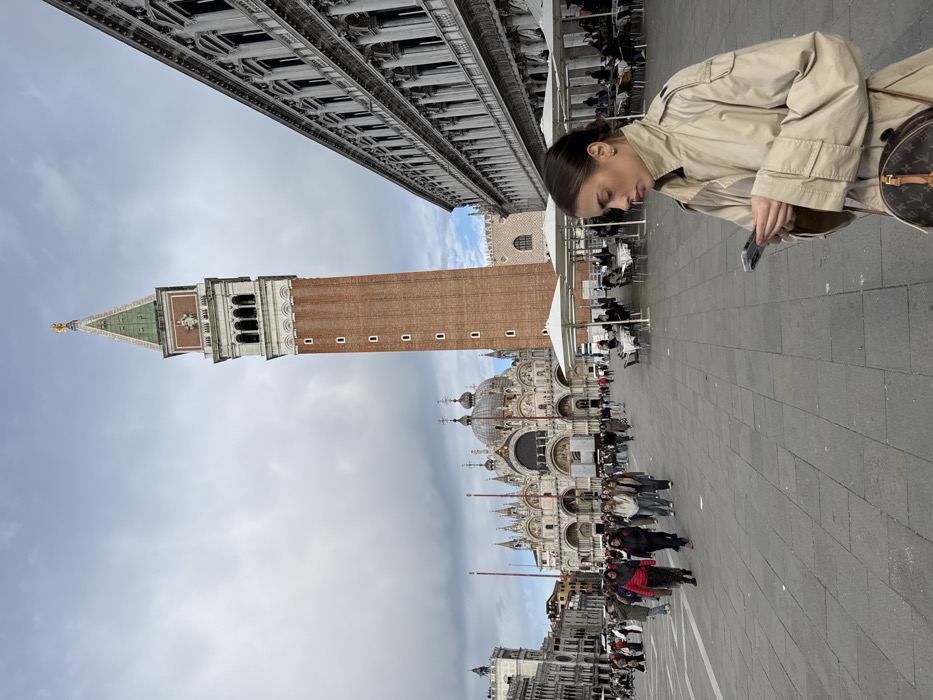 The image captures a wide view of St. Mark's Square (Piazza San Marco) in Venice, Italy, under an overcast sky. In the lower right foreground, a young adult female with brown hair pulled back is depicted in profile, wearing a beige trench coat and carrying a round, brown handbag with a distinctive monogram pattern. She is intently focused on and operating a smartphone held in both hands.
The prominent landmarks of St. Mark's Square dominate the background and mid-ground. The tall, brick St. Mark's Campanile, topped with a green and gold spire, stands centrally. To the left of the Campanile, the ornate, multi-domed facade of St. Mark's Basilica is clearly visible. On the right side of the square, an elongated classical building featuring multiple arched windows and columns (Procuratie Nuove) extends into the background. In front of this building, an outdoor cafe area is arranged with several white umbrellas, tables, and chairs, some of which are occupied by patrons. Numerous individuals are scattered across the large, paved stone square, walking in various directions, indicating a busy public space.