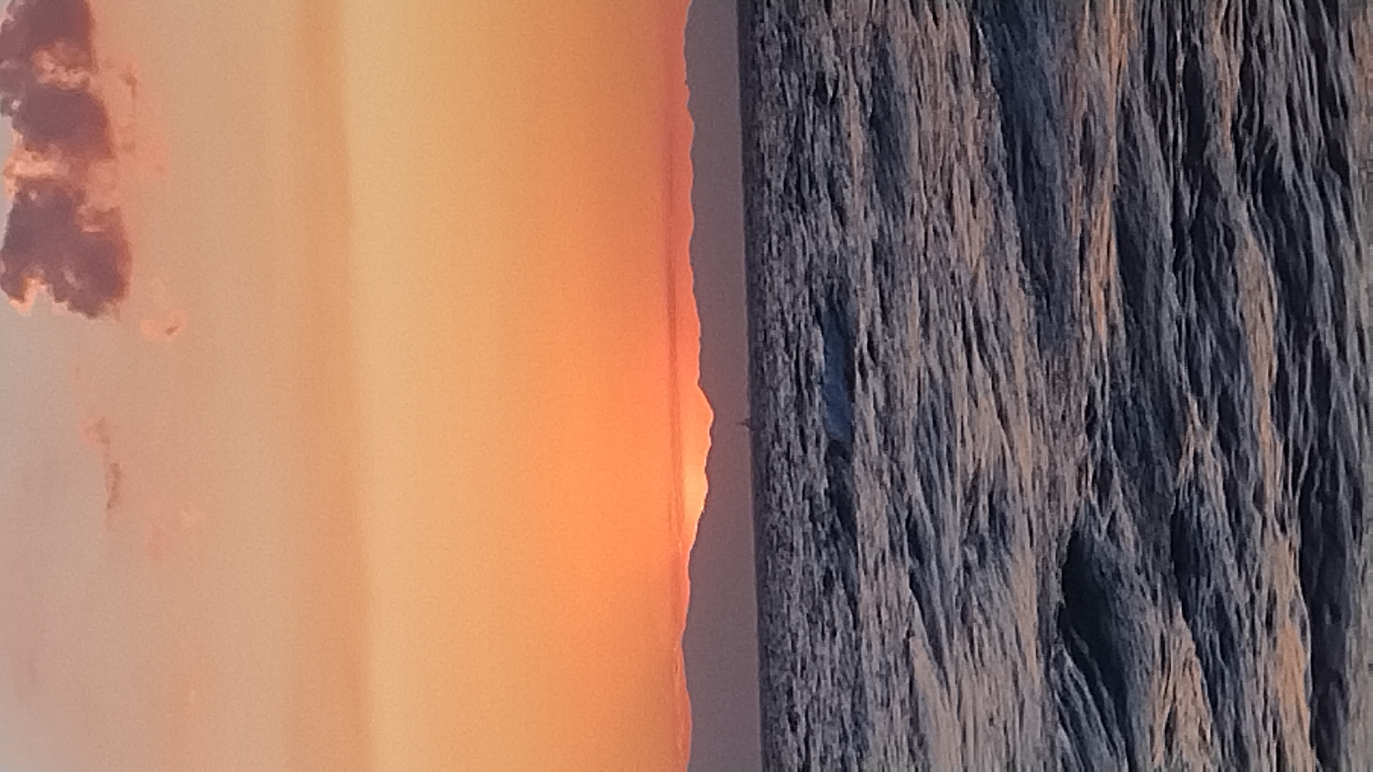 The image depicts a serene seascape at sunset, likely near Neoi Epivates, Greece. The horizon is dominated by a range of silhouetted mountains, behind which the sun is setting, casting a warm, orange glow across the sky. A few scattered clouds in shades of pink and purple are visible in the upper portion of the frame. The sea in the foreground is a deep, dark blue, with gentle waves creating a textured surface. A small, dark object, possibly a sailboat or buoy, is visible on the water in the distance, near the mountains. The overall impression is one of tranquility and natural beauty, typical of a coastal sunset.