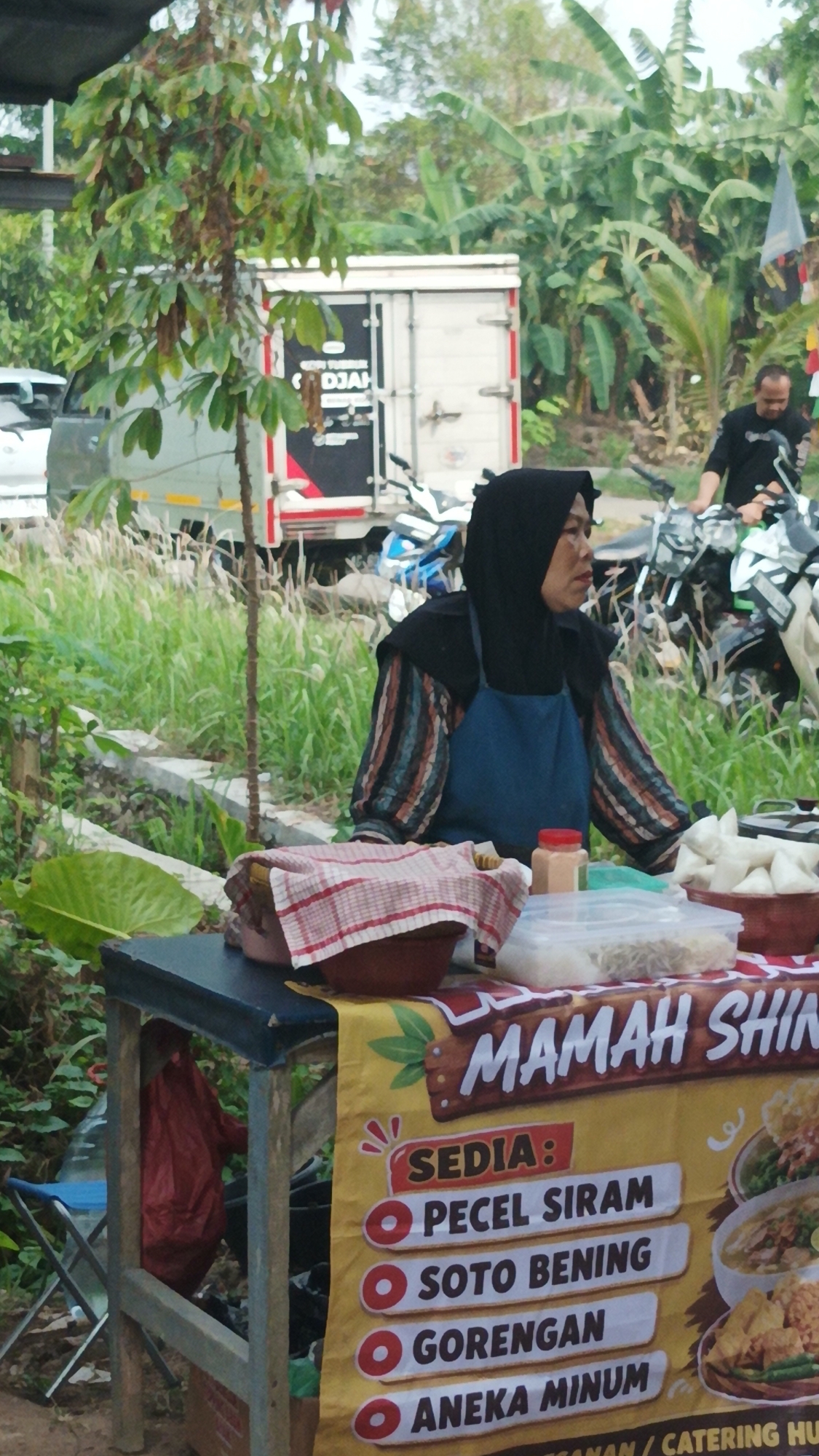 In Munjul, Indonesia, a woman wearing a black hijab and a blue apron stands behind a food stall. The stall features a yellow banner with red text that reads "MAMAH SHIN" and lists "SEDIA: PECEL SIRAM, SOTO BENING, GORENGAN, ANEKA MINUM". In front of her on the stall are various food items in bowls and containers, including rice cakes and what appears to be a dish of fried food. In the background, a white truck with "DJAH" written on it is parked, along with several motorcycles. The environment is outdoors with abundant greenery, including trees and tall grass.