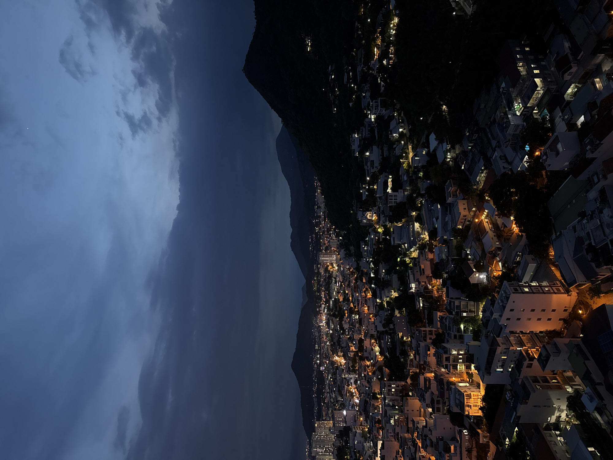 The image shows a twilight aerial view of Bac Nha Trang Ward, Vietnam. The scene is dominated by the transition from day to night, with the sky a deep, cloudy blue, and the city below illuminated by artificial lights. The city is nestled at the base of dark, silhouetted mountains, with the urban sprawl extending towards the foreground. Buildings of various heights and styles are visible, many with glowing windows indicating occupancy. Some taller buildings, likely apartment blocks or hotels, stand out against the lower-rise residential and commercial structures. Streets are traced by lines of light, and the overall impression is of a vibrant, active city settling into its nighttime rhythm. The natural landscape of mountains contrasts with the man-made environment of the city. There are no people visible in the image.