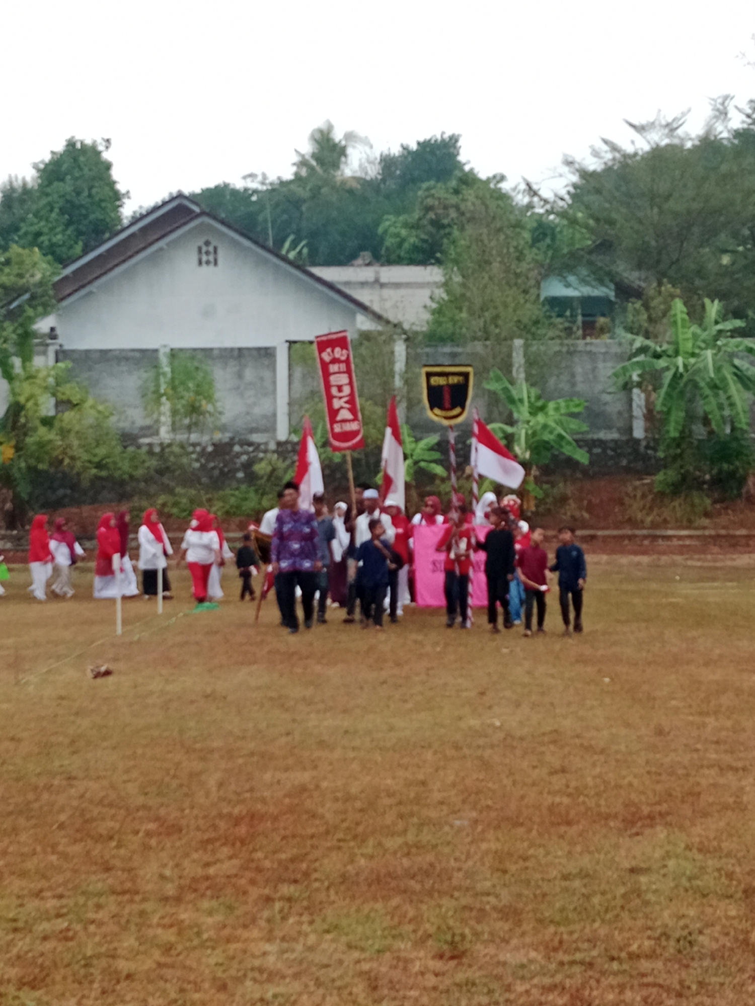 In Bojongmengger, Indonesia, on a dry, grassy field, a group of people are participating in an outdoor event, possibly a parade or a school activity. A white building with a brown roof is visible in the background, flanked by trees and vegetation. The group consists of adults and children, many of whom are wearing red and white attire, with some carrying flags and banners. A prominent red banner with white lettering is held aloft by a man in the center of the image. Other individuals are walking, some carrying what appear to be instruments or props. The atmosphere seems festive, with a sense of community gathering.