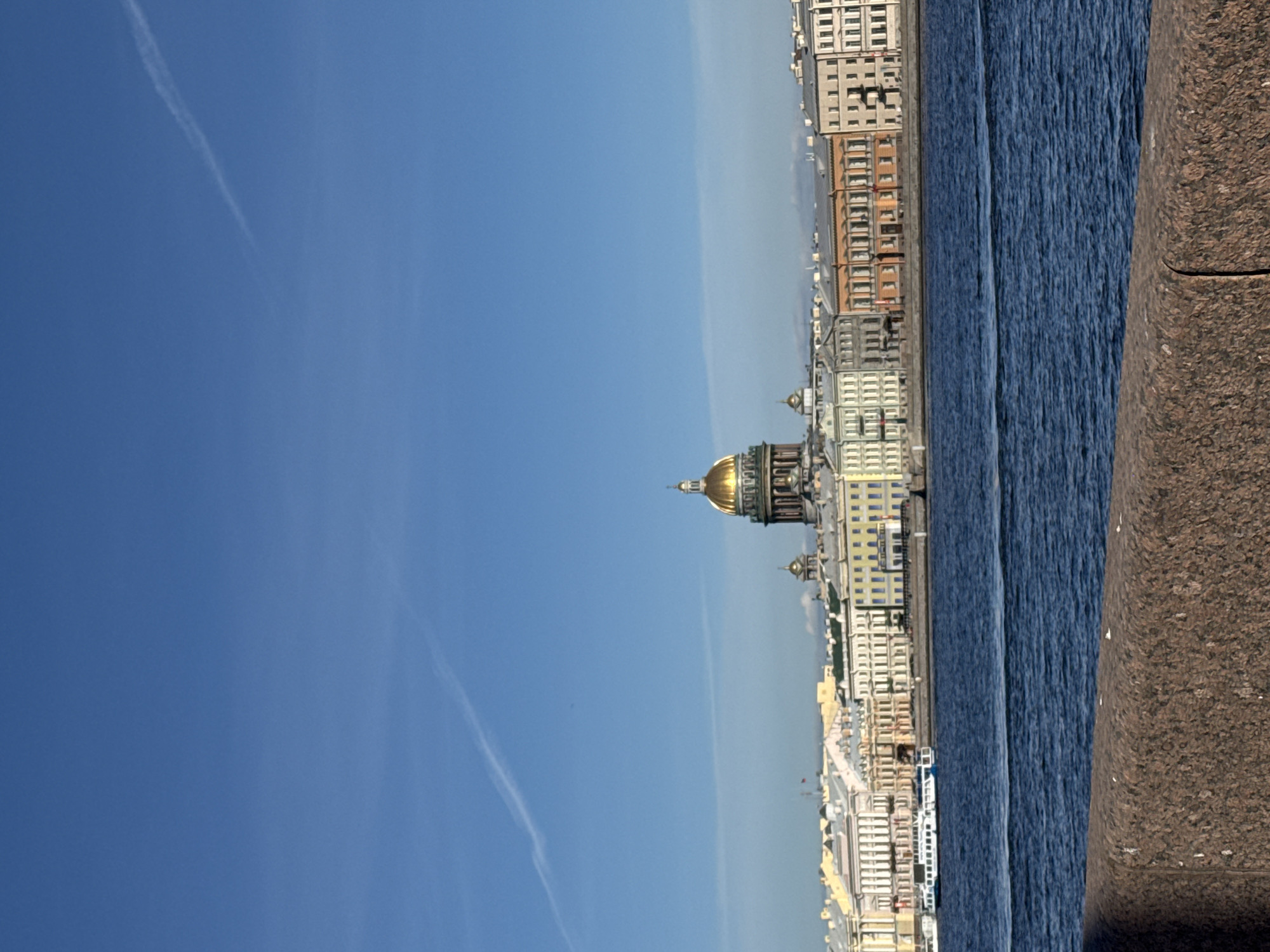 A panoramic landscape view captures a serene cityscape along a body of water in Saint Petersburg, Russia, under a clear blue sky. The image appears to be taken from an embankment, looking across a wide river or canal towards the city's architectural skyline.
In the immediate foreground, a robust granite embankment, reddish-brown and textured, forms a barrier on the bottom of the frame. Beyond it, a expanse of dark blue water, likely the Neva River, stretches across the mid-ground, showing distinct ripples and texture on its surface.
On the far bank of the water, a long line of historic buildings defines the urban horizon. These structures exhibit classical architectural styles, featuring multiple stories, numerous windows, and facades in varying shades of cream, yellow, and light brown. Dominating the left side of this skyline is the iconic golden dome of St. Isaac's Cathedral, brightly gleaming under the sunlight and standing prominently above the surrounding buildings. Other smaller domes or architectural features are also visible among the building complex.
The background is dominated by a vast, clear blue sky, transitioning to a lighter, hazier blue closer to the horizon. A single, faint white contrail trails diagonally across the upper left portion of the sky. The time of day is bright daylight, suggesting a sunny morning or afternoon. No people or specific activities are visible, presenting a calm and grand vista of the city. There is no discernible text in the image.