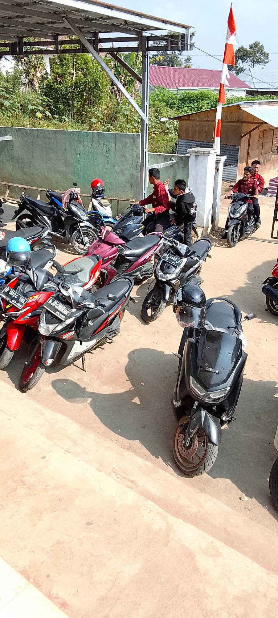 This is a medium-angle, eye-level shot of a collection of parked scooters and motorcycles outside a building in Jepara, Indonesia. The scene appears to be a sunny day, with strong shadows cast on the light-colored ground. Several people are present, some sitting on the scooters, and others standing. The subjects are predominantly young males, dressed in casual clothing, with some wearing red shirts or jackets.
In the foreground, a black scooter is parked, with a black helmet resting on its handlebars. To its left, a red and black scooter is visible, followed by a dark red scooter and a black and blue scooter. Several other scooters are parked behind these, creating a dense arrangement.
In the mid-ground, a group of young men are gathered. One man in a red shirt is sitting on a black scooter, gesturing with his hand. Another young man in a black hoodie is leaning on the same scooter, looking towards the man in red. Further to the right, two young men in red shirts are on another black scooter, with one appearing to be driving.
In the background, a building with a corrugated metal roof is visible. A flagpole with an Indonesian flag is positioned to the right of the frame, with the red and white flag fluttering in the breeze. Lush green vegetation surrounds the building, with trees and bushes clearly discernible. The overall impression is of a casual gathering or a place where people are parking their vehicles before entering the building.
