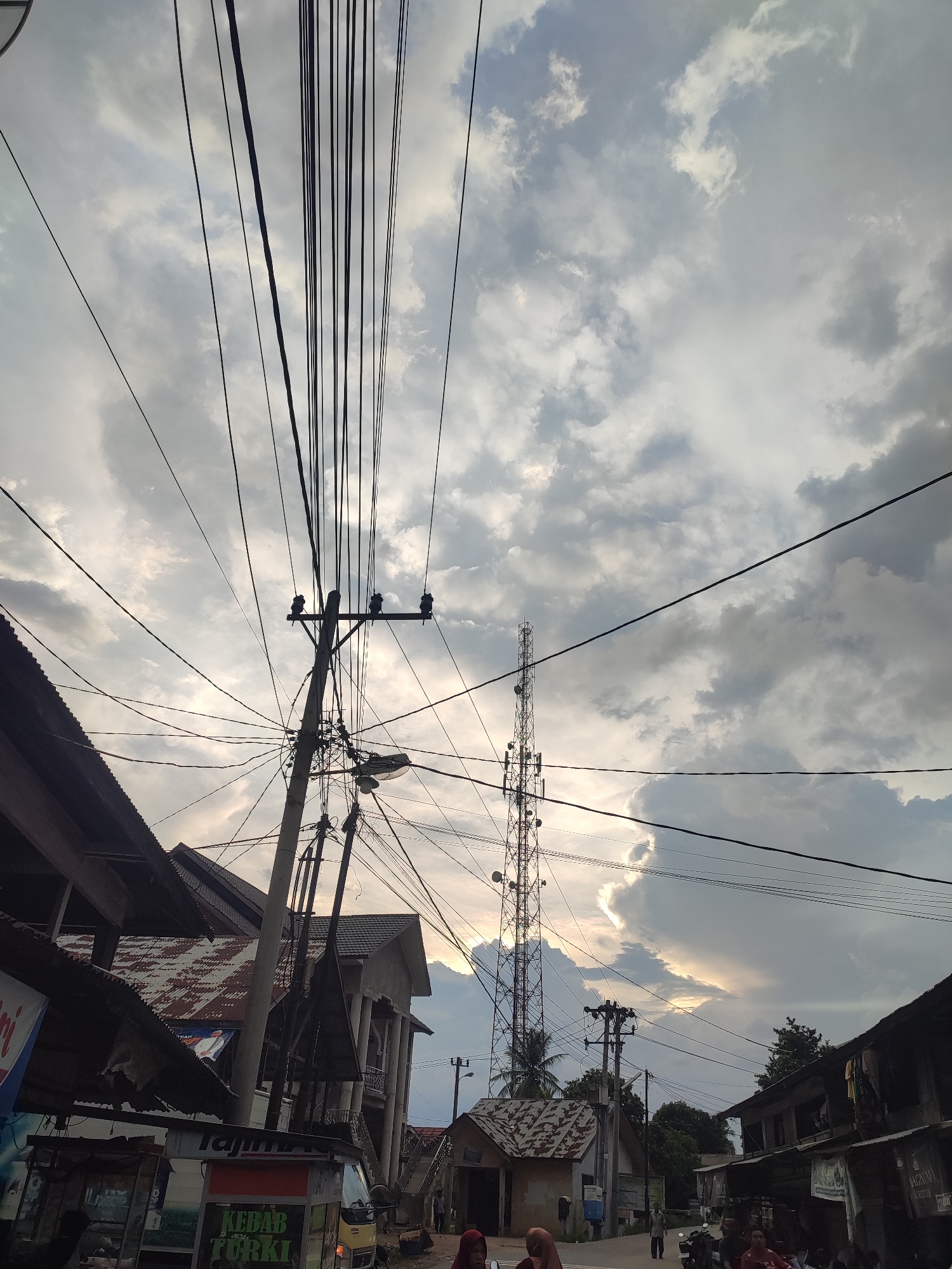 The image shows a street scene in Pantonrayeuk Satu, Indonesia. The sky is filled with gray and white clouds, with a hint of sunlight breaking through. A tall communications tower is visible in the middle distance, surrounded by a network of power lines and electrical poles. The foreground includes buildings with corrugated roofs. A kebab stand is visible on the left side of the image. Several people are present on the street.
