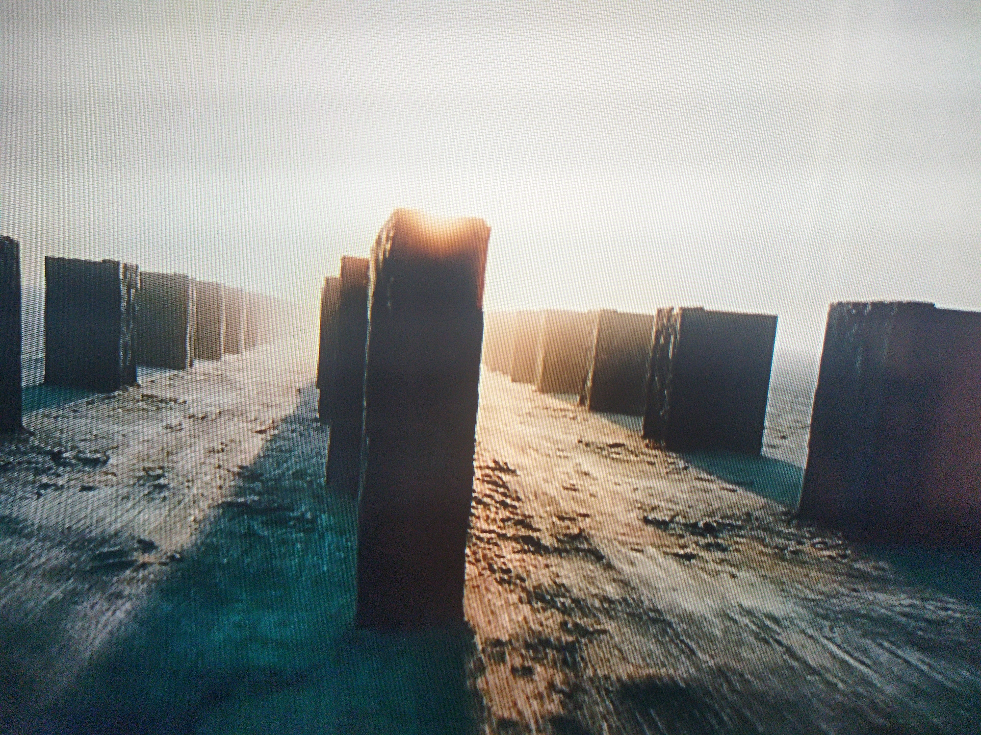The image shows a landscape with a series of dark, rectangular structures arranged in a linear formation. The ground appears textured and uneven, with a combination of blue, brown, and grey tones. The sky is bright and hazy, with the sun partially obscured behind one of the structures, creating a backlighting effect. The overall scene suggests a desolate or industrial setting, but there is no visible information or evidence to indicate the location in Kramatorsk, Ukraine, or any other specific location. There are no people visible in the image.