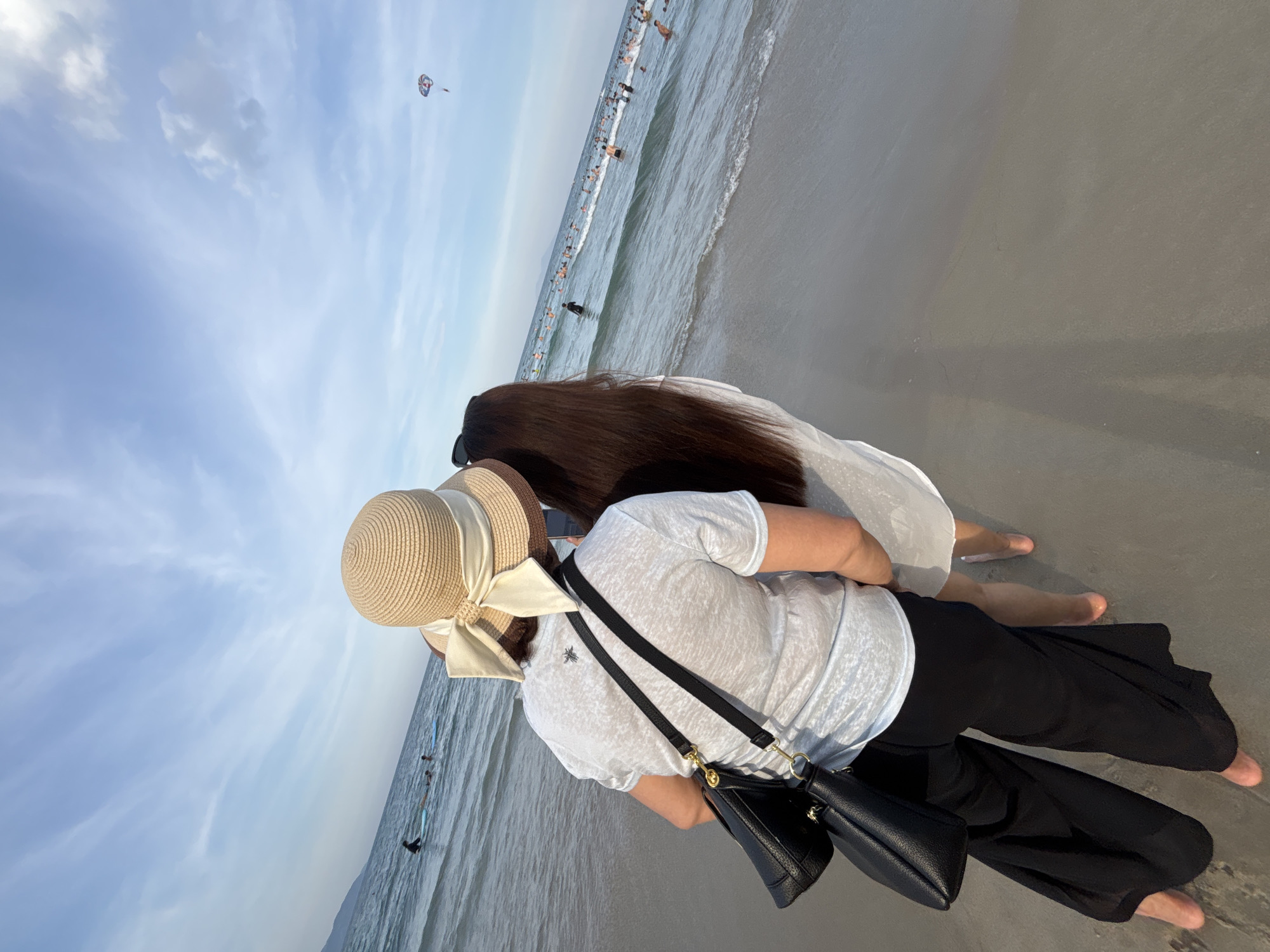 The image shows two women standing on a sandy beach, facing the ocean. The woman in the foreground is wearing a white t-shirt, black pants, and a straw hat with a large bow. She has a black purse slung over her shoulder. Her hair is long and dark, and she is wearing sunglasses. The woman behind her is wearing a white dress and also has long dark hair.
The ocean is calm with small waves, and many people are in the water, swimming and enjoying themselves. In the distance, a parasailer is visible in the sky. The sky is a pale blue with some scattered clouds. The sun appears to be low in the sky, suggesting it might be late afternoon or early morning. The scene is lively, with a sense of people enjoying a day at the beach. The image is oriented vertically, with the water appearing to be at the top and the sand at the bottom. The context of Da Nang, Vietnam, places this scene in a popular tourist destination known for its beaches.