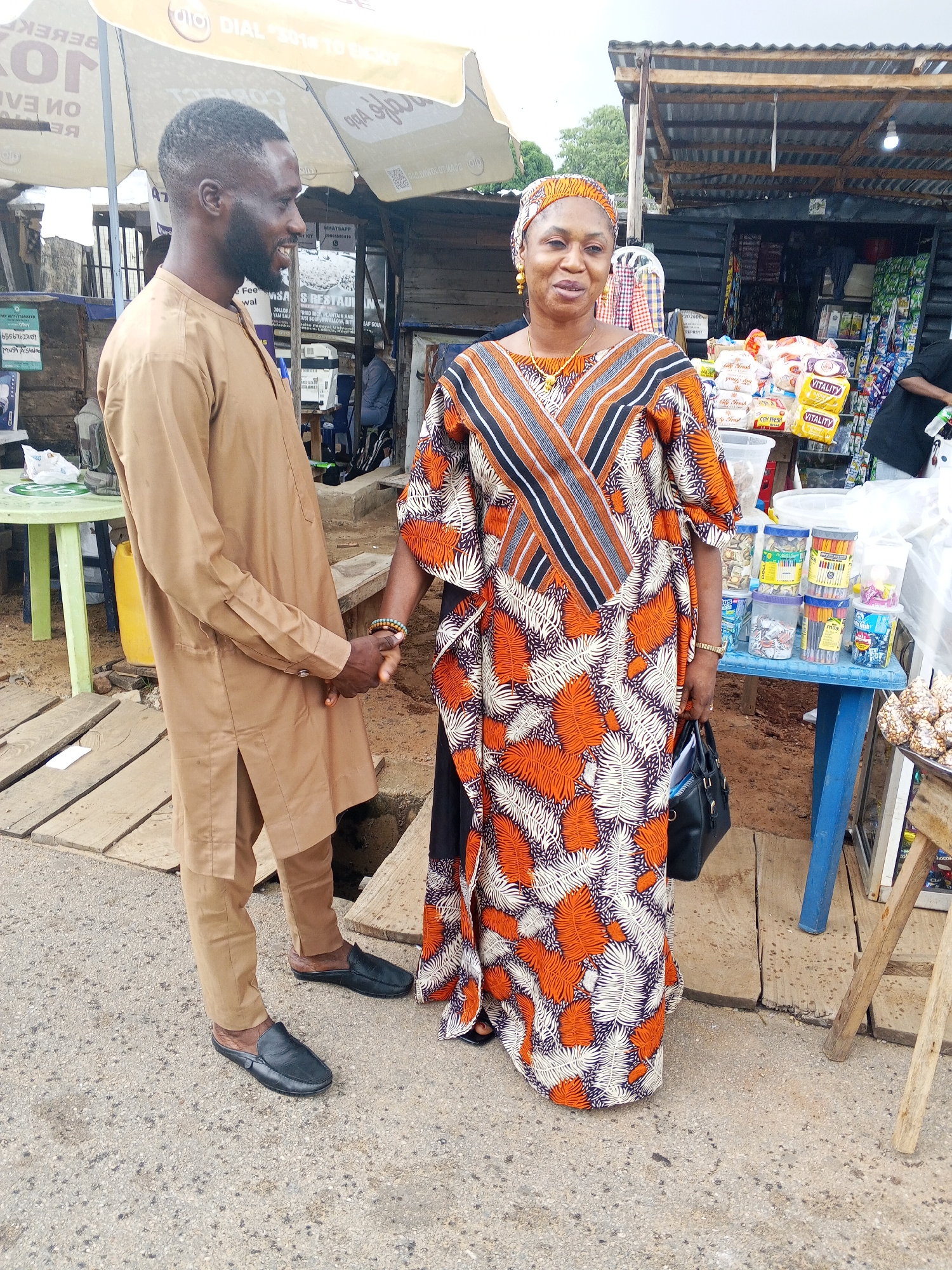An African man and woman stand on a paved area with wooden planks in the foreground, engaging in a handshake. The man, wearing a tan traditional outfit, faces left, smiling. The woman, in a vibrant orange, white, and black patterned dress and headscarf, faces forward, holding a black handbag. Behind them, a market stall is visible, stocked with various goods. The environment appears to be an outdoor market in Lokoja, Nigeria, with a cloudy sky overhead.