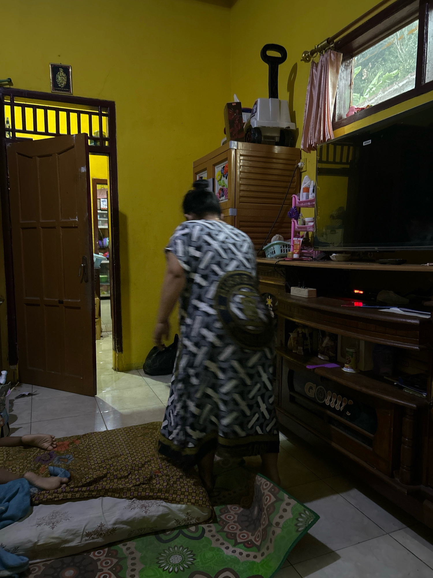 The image depicts an indoor scene in a room with yellow walls. A woman wearing a black and white patterned dress is standing and facing away from the camera, walking towards the right of the frame. In the foreground, on the floor, is a person lying down on a patterned mat, with their feet visible and covered by a blue blanket. To the left of the woman, a wooden door is slightly ajar, revealing a hallway with another open door and shelves filled with items. In the background, to the right, a large television is mounted on a wooden entertainment unit, and a window with pink curtains is visible above it. Various items, including a white toy truck with a black handle, a pink shelf with containers, and a black bag, are scattered around the room. The overall impression is of a lived-in domestic space. The provided text indicates the location is Cihaurbeuti, Indonesia, suggesting this is a residential setting within that area.