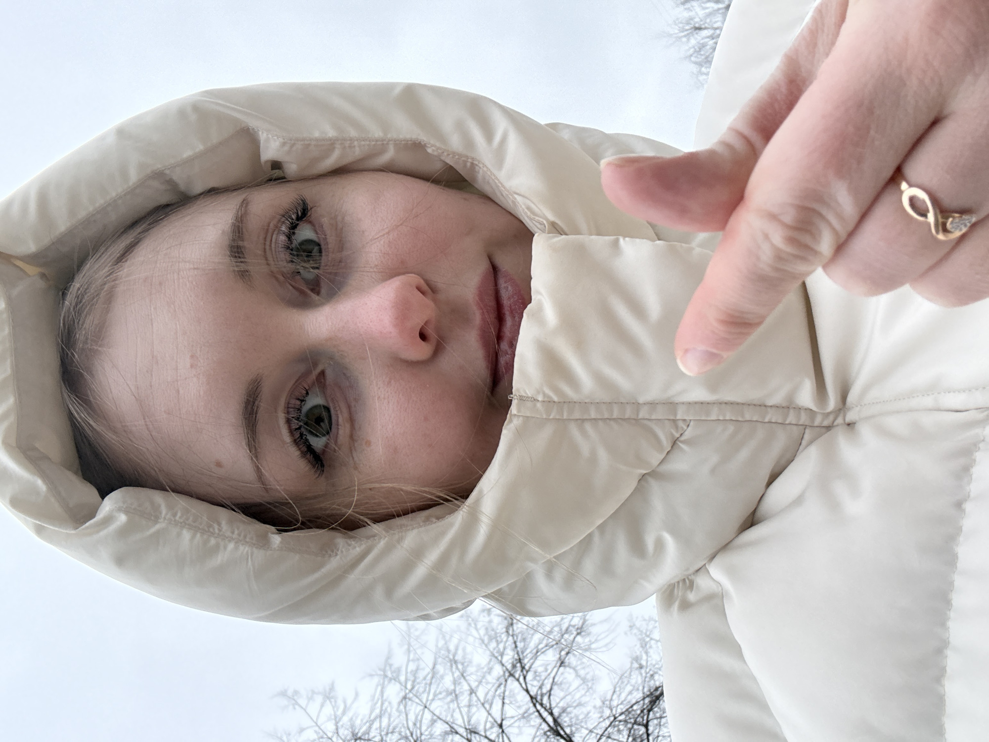 A close-up, upward-angled portrait shows a fair-skinned person with light brown hair in a cream-colored hooded jacket against a pale sky. Their face, featuring green eyes, is angled slightly. A hand with a gold ring on its ring finger points toward the person's face from the right. Thin, leafless tree branches are visible in the background, suggesting a winter setting. The location is Unknown, Unknown.