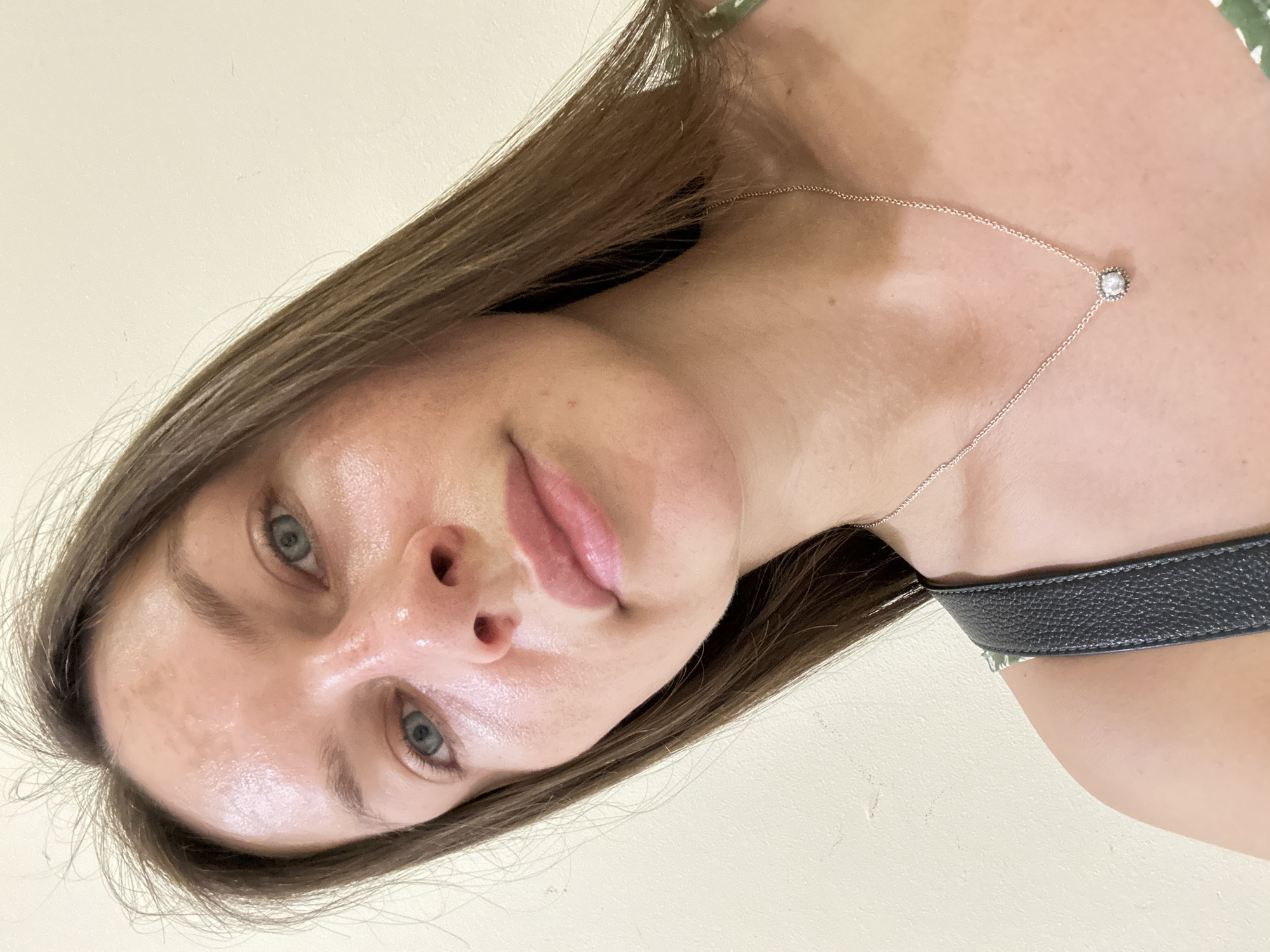 A woman is captured in a close-up selfie, her head and shoulders dominating the frame. She has long, brown hair that falls past her shoulders and light blue eyes. Her skin appears to have a dewy, possibly oily, sheen. She is wearing a thin silver necklace with a small, square pendant. A dark strap, likely from a bag or clothing, is visible across her left shoulder and chest. The background is a plain, light-colored wall, suggesting an indoor setting. There is no indication of the city of Budva, Montenegro, in the image.