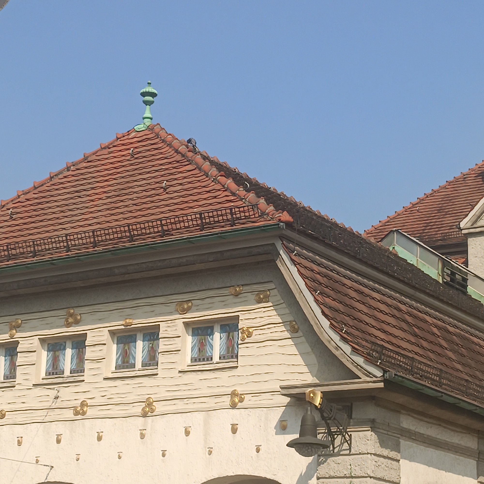 The image shows the facade of the Sprudelhof in Bad Nauheim, Germany, a striking example of Art Nouveau architecture. The building features a red-tiled roof with a decorative green finial at its peak. The facade itself is a light beige color, adorned with horizontal decorative lines and numerous gold circular ornaments. Three distinct windows with stained glass are visible, each framed by the decorative elements. A black bell hangs from a decorative metal bracket on the right side of the building. The sky above is clear and blue, indicating fair weather.