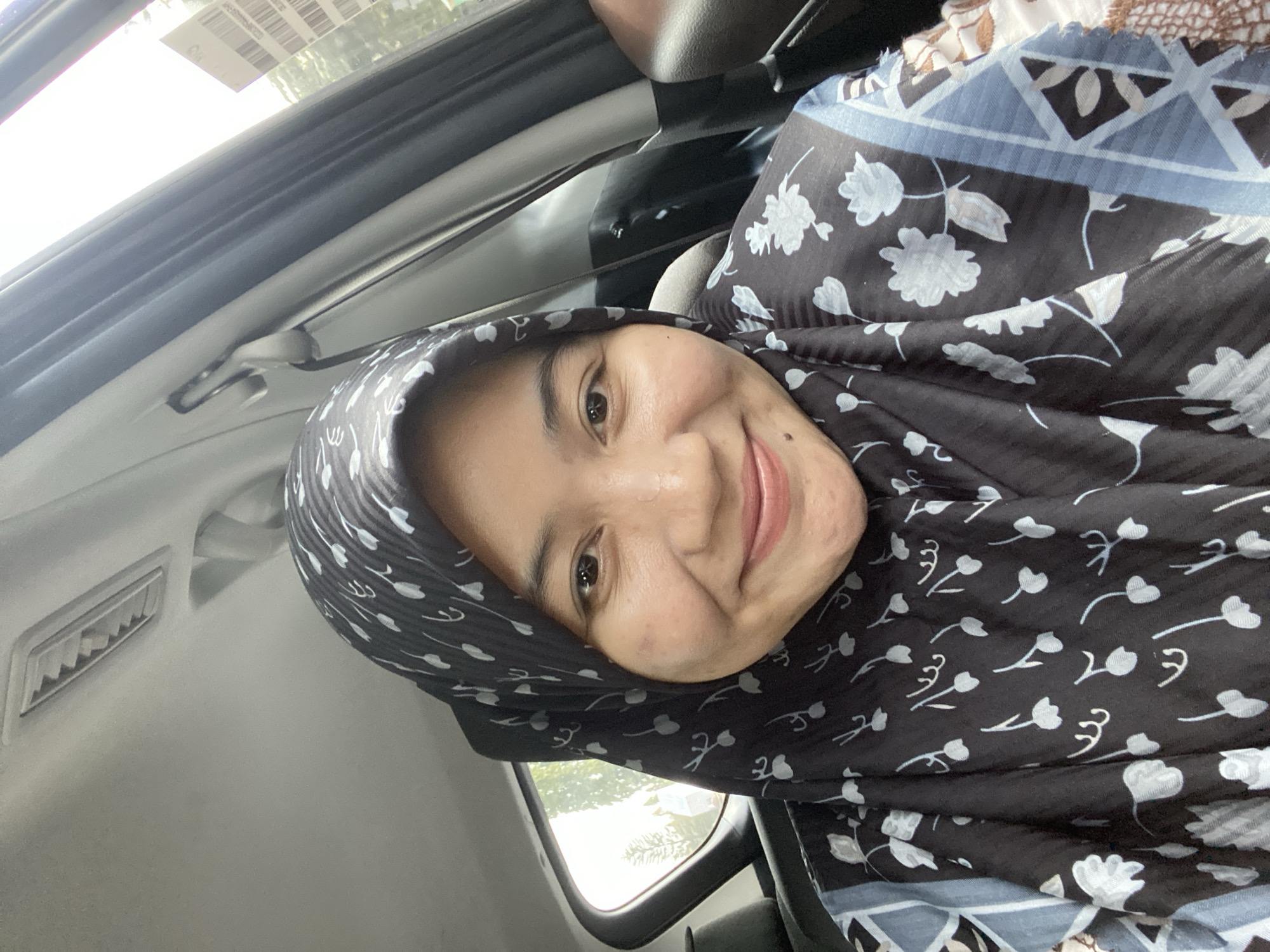 A medium shot captures a young woman with light brown skin and dark brown eyes, wearing a black hijab with a white floral pattern. She is positioned in the passenger seat of a car, looking directly at the camera with a slight smile. The car's interior is visible, including the ceiling light, a seatbelt, and the window. Outside the window, trees and a glimpse of buildings suggest an urban setting, possibly in Banjarbaru, Indonesia, given the context. The overall lighting suggests it is daytime.