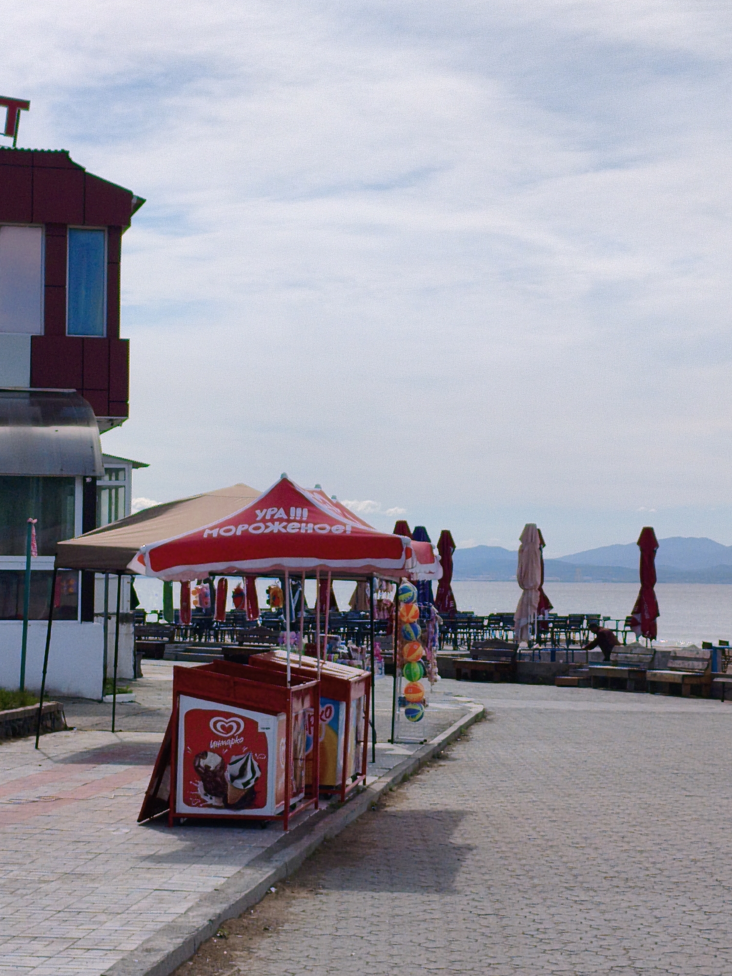 The image shows a paved walkway along a waterfront area in Unknown, Unknown. On the left is a building with a reddish-brown facade and a window. A covered patio extends from the building, supported by white posts. In the foreground, there are red ice cream vendor carts with advertising for ice cream. A red-and-white umbrella covers the carts, bearing text in a foreign language (Russian) "УРА!!! Мороженое!", meaning "Hooray!!! Ice cream!". Hanging from the umbrella are toys or small items for sale.
In the background, there's an outdoor seating area with tables and chairs, protected by closed umbrellas. Further back, the sea or a large lake is visible, with mountains or hills in the distance under a sky with scattered clouds. A single person can be seen near the water, appearing to be bending over or working. The scene suggests a tourist or recreational area.