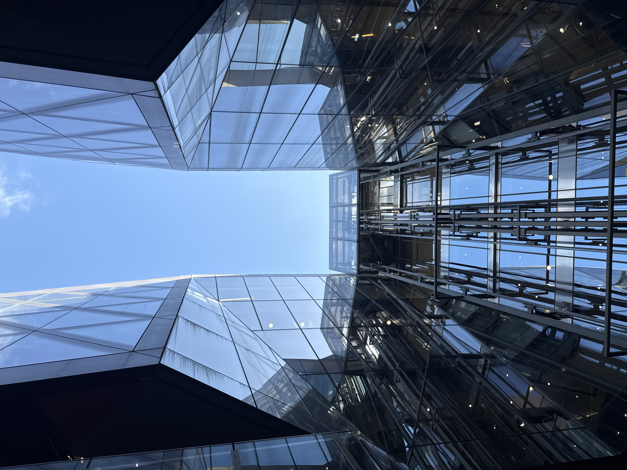 The image shows a low-angle view looking up at modern architecture. Two buildings with glass facades are visible, angled toward each other with a narrow gap of clear blue sky between them. The building on the right is intricate, with an open elevator shaft. The building on the left features triangular glass panels. The location appears to be in the city of London, United Kingdom.