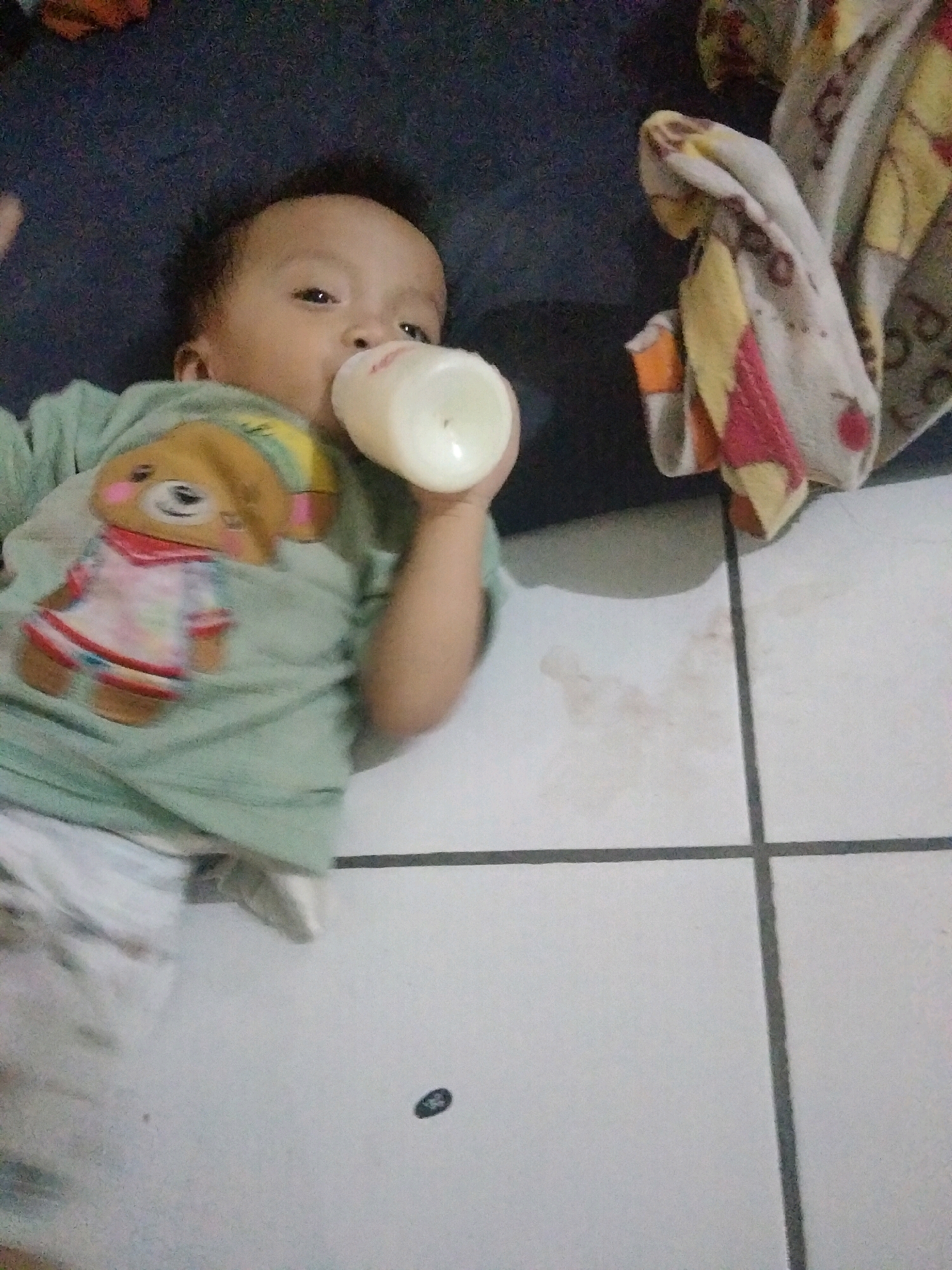 A baby is lying on its back, holding a baby bottle. The baby is wearing a light green t-shirt with a teddy bear graphic on the front. To the right of the baby, a folded blanket with a colorful pattern is visible. The background consists of a dark surface, possibly a bed or couch, and a tiled floor with some light brown stains. The scene appears to be indoors, in a residential setting in Suka Sari, Indonesia.