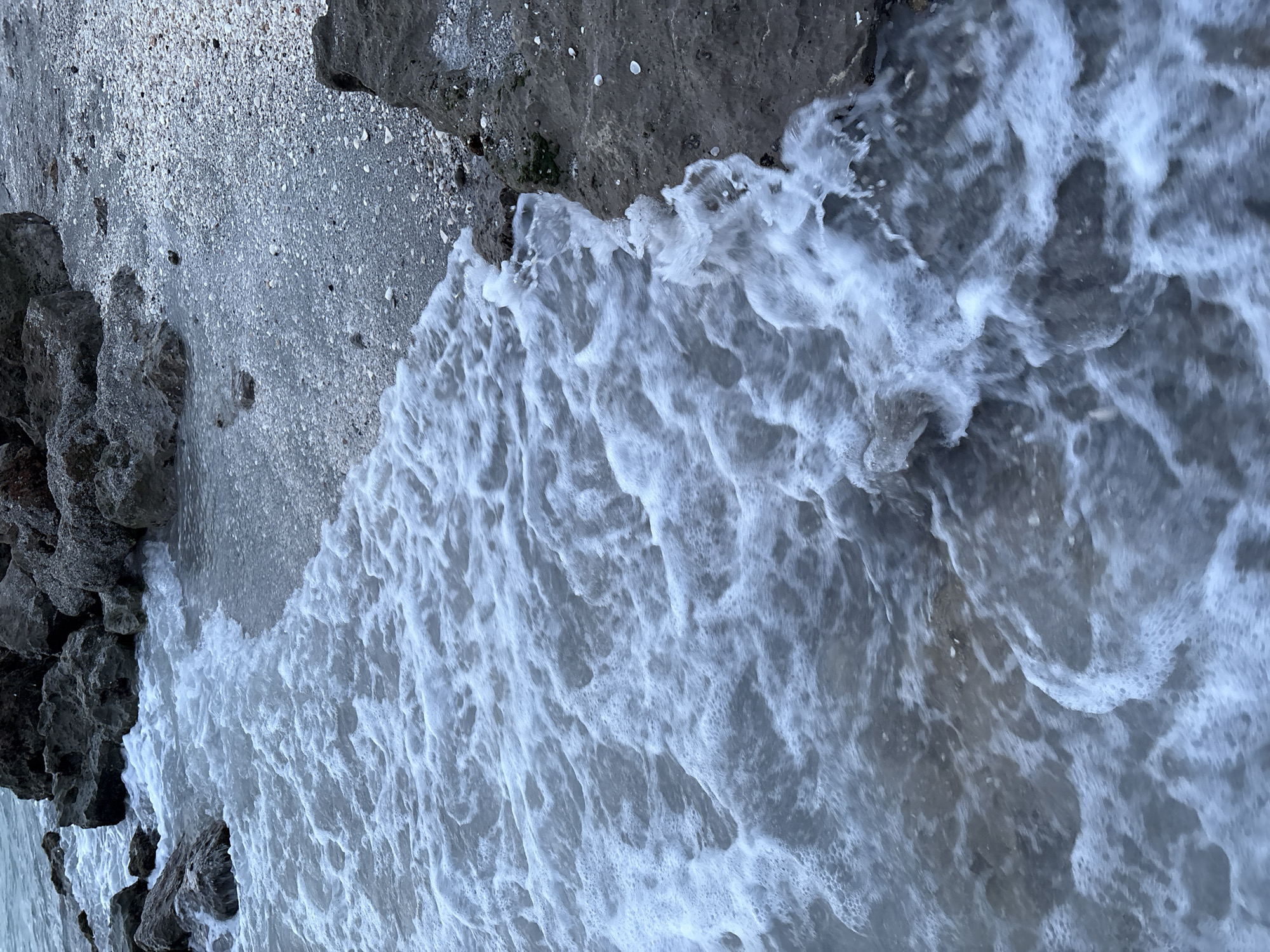 The image shows a close-up view of waves gently lapping against a rocky shoreline covered in shells and small stones. The water is foamy and appears to be moving from the left to the right side of the frame, interacting with the dark, textured rocks and the pebble-strewn beach. The overall tone of the image is muted and natural, suggesting an outdoor, coastal environment. The scene is devoid of any human presence or artificial elements. The location is identified as Venice, United States of America, which implies a beach or coastal park setting within that city.