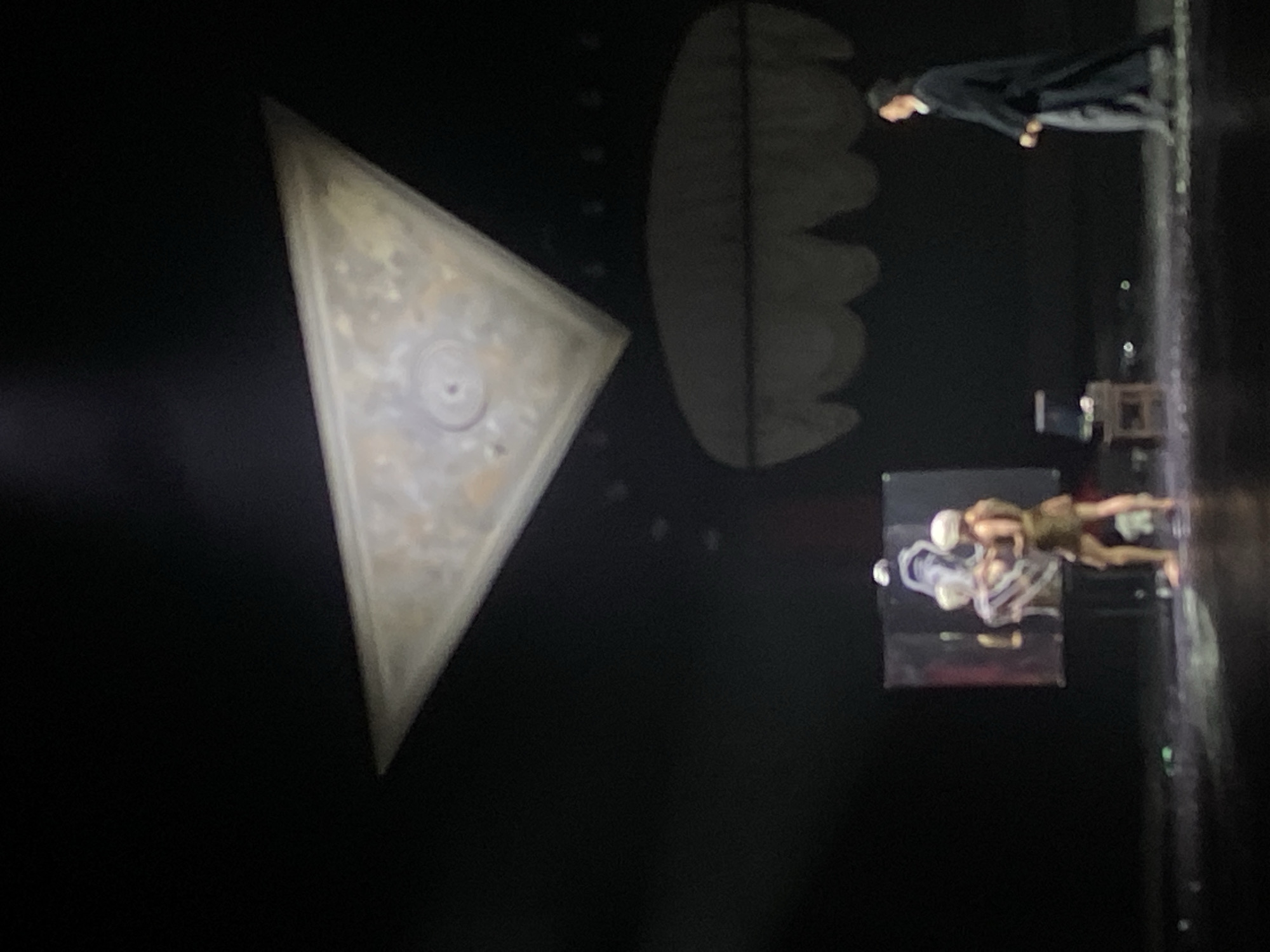The image shows a performance on a stage in Заволжье, Russia. There are two performers interacting in the lower right, wearing neutral-toned clothing. Behind them is a screen displaying what appears to be a medical or anatomical image with bright white, somewhat bulbous shapes. To the left, hanging from the ceiling, is a large triangular object with a design. To the right of the triangle is a curved object that looks like a large comb. A person in a dark coat stands near the stage edge in the upper right. A row of lights is visible on the edge of the stage. The background is dark and undefined.
