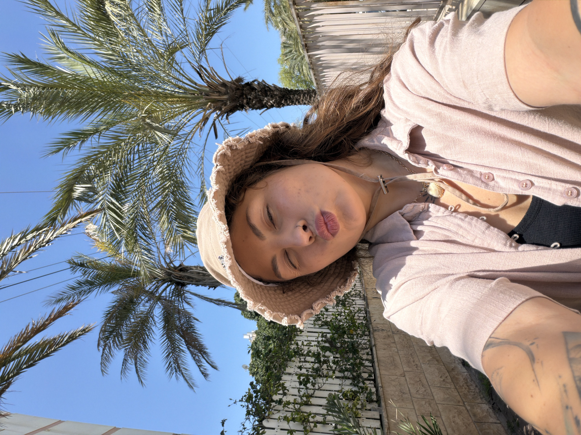 A young person takes a close-up selfie outdoors on a bright, sunny day in Kiryat Bialik, Israel. The individual, with long, dark brown hair, is wearing a light pink or beige bucket hat and a light pink long-sleeved button-up shirt over a black top. Their eyes are closed, and their lips are playfully pursed in a "kiss" expression. Two necklaces adorn their neck: one featuring a seashell pendant on a cord, and another silver chain with a t-bar clasp. A tattoo is partially visible on their right arm.
The background is bathed in strong sunlight, suggesting midday or early afternoon. A clear, vibrant blue sky fills the upper portion of the frame, dotted with several large palm trees whose green fronds extend into view. Below the sky and palm trees, a white slatted fence is visible, covered with lush green foliage, possibly ivy. The ground or a low wall appears to be made of light-colored stone or tiles. The overall atmosphere is bright, warm, and relaxed. No visible text is present in the image.