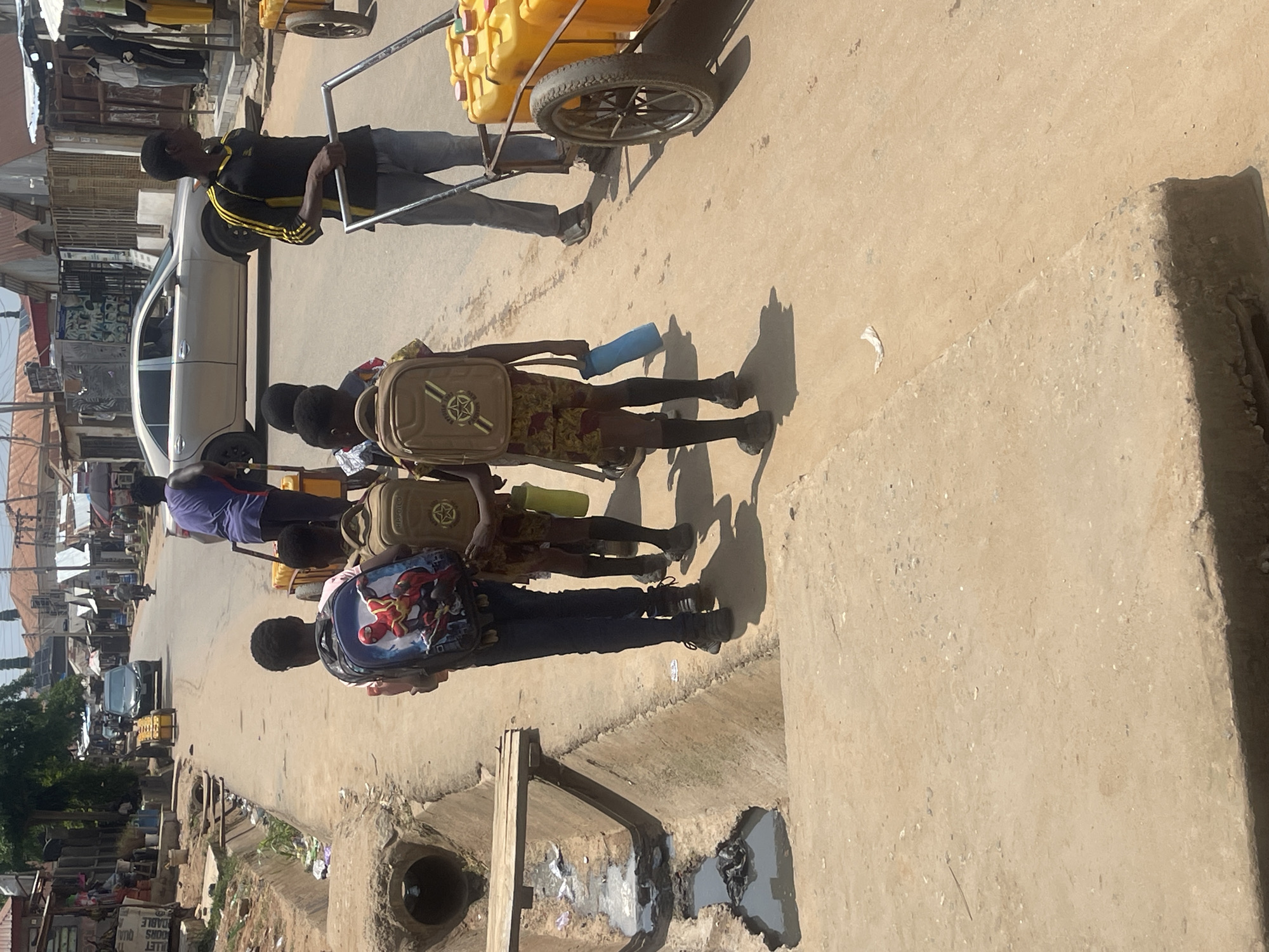 In the city of Abuja, Nigeria, a group of individuals are seen walking along a dirt road lined with buildings and a drainage ditch. In the foreground, three young males are walking away from the camera. The boy on the left wears a Spider-Man themed backpack and dark pants. The boy in the middle has a backpack with a star emblem and is wearing patterned shorts. The boy on the right also wears a similar backpack and patterned shorts. To the right of the children, an adult male in a black and yellow shirt is pushing a cart loaded with several yellow jerrycans. In the background, a silver sedan is driving down the road, and other people and structures are visible. The environment appears to be a busy street with evidence of commerce and daily life. The drainage ditch in the foreground contains murky water and some debris. The lighting suggests it is daytime, likely mid-morning or afternoon, with strong sunlight casting shadows.