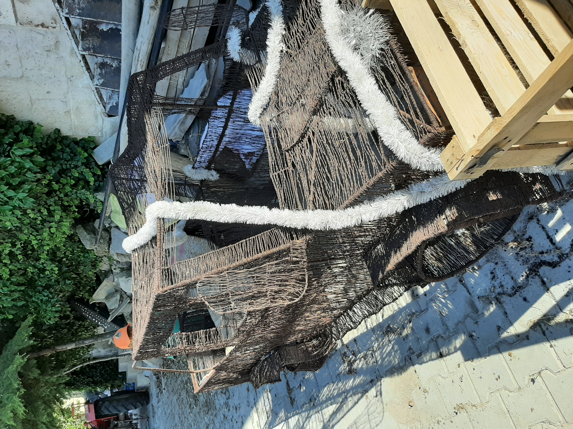 In Uçhisar Beldesi, Turkey, a wire-frame sculpture resembling a car sits outdoors on a paved surface. The sculpture is brown and appears to be made of woven wire. White, fluffy trim is attached to the top and sides of the car sculpture. To the left of the sculpture, a red tractor is partially visible. The background includes lush green foliage, a stone wall, and various discarded materials, including wooden crates and metal objects. The scene is illuminated by sunlight, casting shadows on the ground.