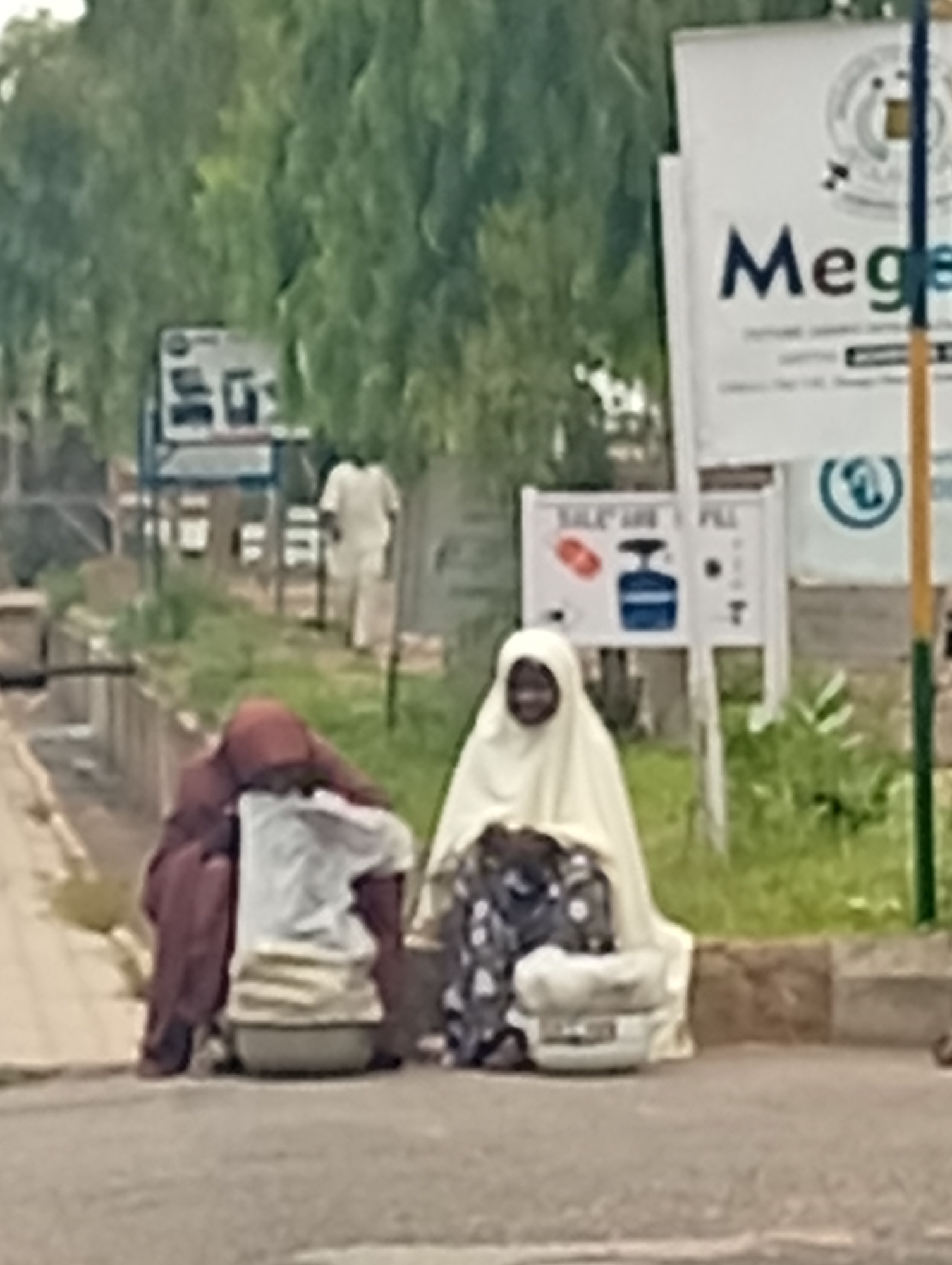In Gombe, Nigeria, two women are seen sitting on the roadside in front of a white sign with "Mege..." visible. The woman on the right wears a cream-colored hijab and a patterned dress, holding a white bundle. The woman on the left is draped in a maroon garment and has a white bundle in front of her. A man in a white outfit walks away in the background. The setting includes lush green trees, a paved road, and a concrete curb. A blue and yellow pole is also visible near the sign.