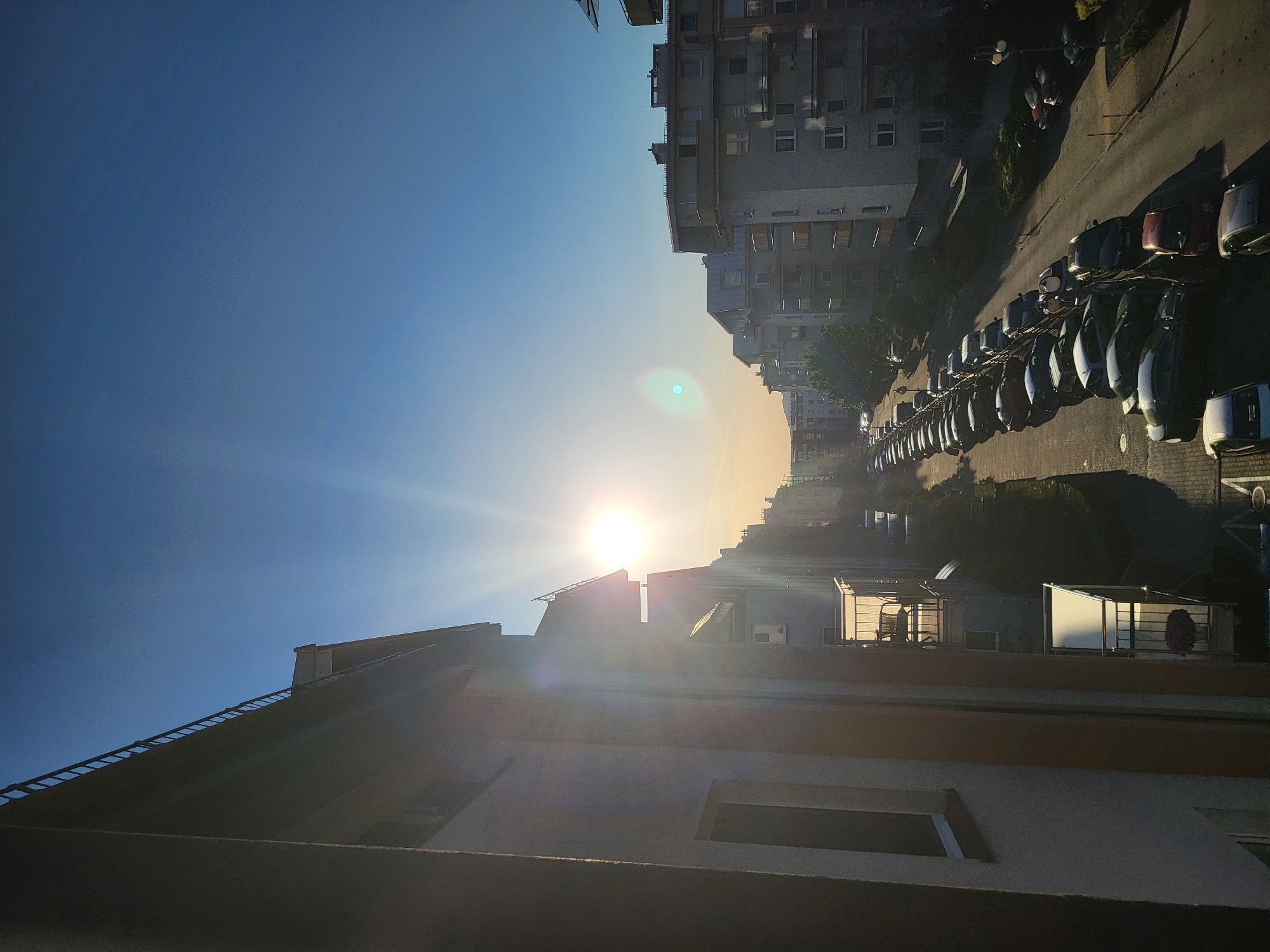 An outdoor, eye-level, vertical shot captures a city street lined with apartment buildings and parked cars under a bright blue sky with the sun shining. The sun, positioned in the upper left quadrant of the frame, casts a strong lens flare across the scene, illuminating the facades of the buildings and creating long shadows on the street. The buildings are multi-story residential structures with visible windows and balconies. A long line of cars, varying in color and model, are parked along the side of the street. The foreground is dominated by the side of a large apartment building. The overall scene suggests a residential neighborhood in a city during daylight hours.