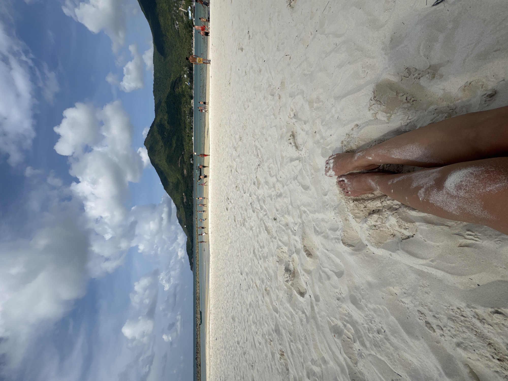 The image depicts a sandy beach in Baan Chaloklum, Thailand. The frame is oriented vertically. The lower right portion shows a pair of bare human legs, covered in sand. The foreground is primarily composed of white sand, revealing footprints and other disturbances. In the midground, a body of water separates the beach from a mountain densely covered with green vegetation. A line of people is visible along the shoreline. Above the mountain, a blue sky is filled with scattered white clouds.
