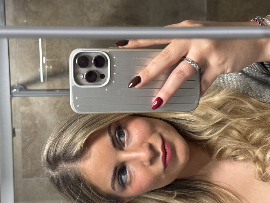 A woman with blonde hair and blue eyes takes a selfie in a mirror. She holds a phone with a silver, rectangular case. Her fingernails are painted in dark red and brown tones. A ring is visible on her ring finger. The background features beige tiles and a chrome towel rack. The overall image is slightly rotated.