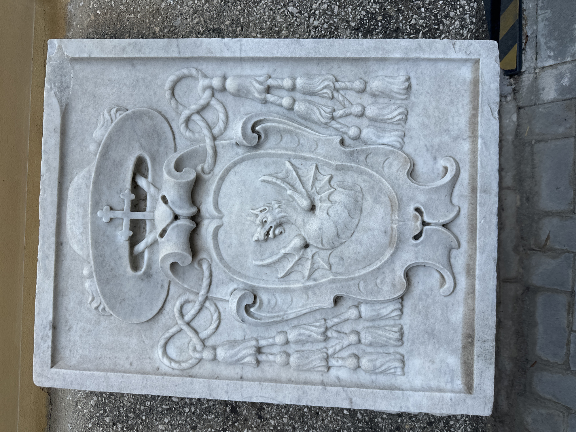 A white marble relief carving of a dragon crest is mounted on a textured wall outdoors. The crest features a dragon with outstretched wings within a shield, flanked by ornate tassels and ropes. Above the shield is a bishop's mitre with a cross. The carving is weathered and appears to be an architectural element or historical marker. The background wall is a pale yellow, and the ground is paved with gray stones. The location is the exterior wall of the Sacristy of the Basilica di San Lorenzo Maggiore in Naples, Italy.