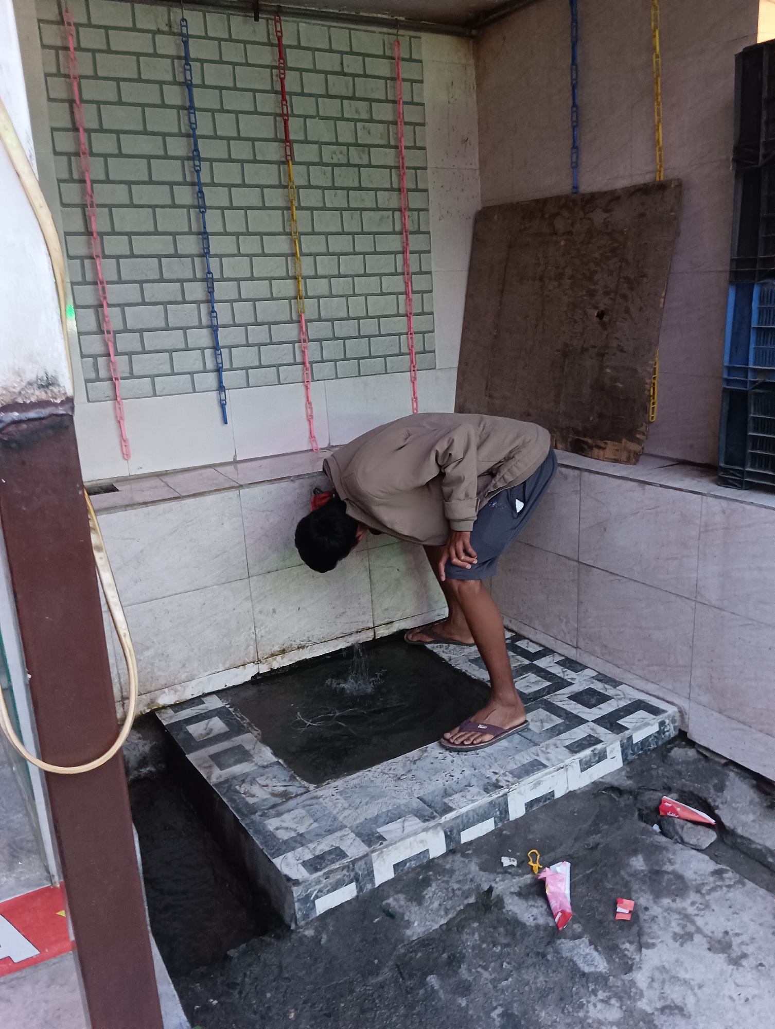 A person is bent over a small, tiled water trough, cupping water to their face to drink. They are wearing a light brown jacket and dark shorts, with brown flip-flops on their feet. The trough is outdoors, with a tiled wall behind it featuring decorative chains in pink, blue, and yellow. To the right of the trough, a large wooden board leans against the wall. The ground around the trough is wet and grimy, with discarded wrappers and a yellow plastic item visible. The overall impression is of a simple, utilitarian space, possibly a public wash area in a warm climate. The lighting suggests it is daytime.