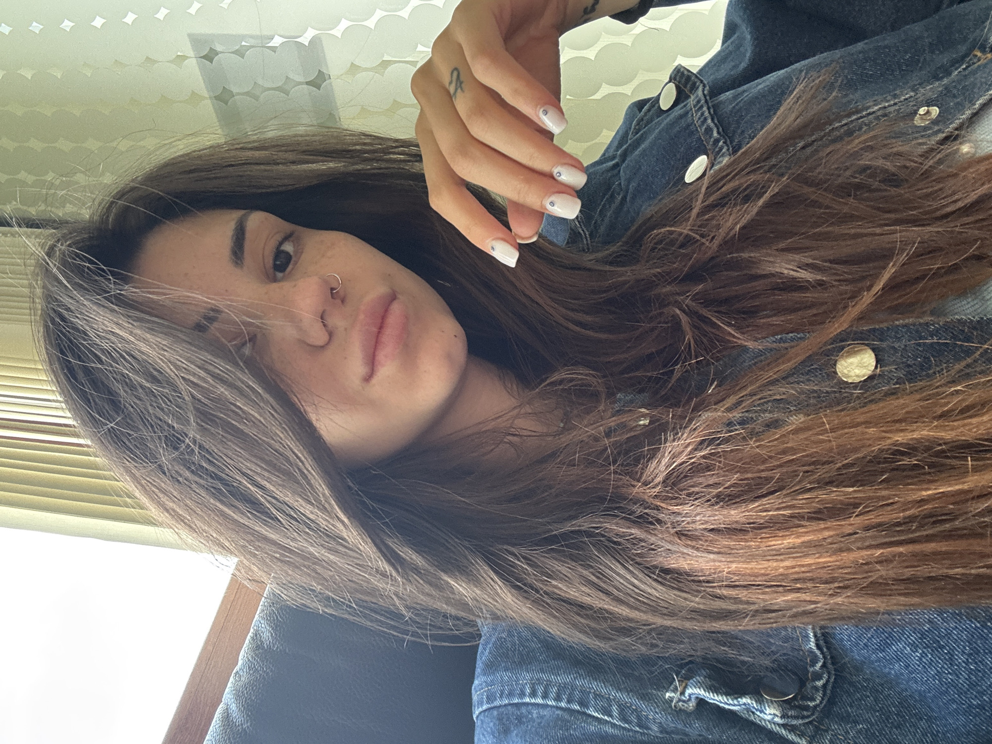 This is a close-up portrait of a young woman, captured in a selfie. She has long, straight to slightly wavy dark brown hair, which frames her face and flows over her shoulders and chest. Her fair skin is visible, adorned with freckles across her nose and cheeks. She has a nose piercing, a small silver hoop in her right nostril, and her lips are slightly parted in a subtle pout.
She is wearing a blue denim jacket with metallic buttons, visible on her shoulders and lapel. Her right hand is raised towards her head, showing off her white manicured nails, which each feature a small, dark circular embellishment. A small, delicate tattoo, possibly a symbol or letter, is visible on the middle finger of her right hand.
The background on the upper left side of the image reveals an indoor setting with a light-colored, patterned panel or ceiling and horizontal venetian blinds below it. The rest of the background is darker and indistinct, suggesting a car interior or dark furniture. Bright, natural light illuminates her, indicating it is daytime. There is no visible text in the image.