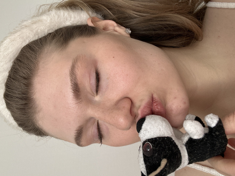 A close-up, front-facing portrait depicts a young adult female from the upper chest upward, performing a kissing gesture towards a small, black and white stuffed animal. The female has light brown hair, pulled back by a fluffy white headband. Her eyes are closed, and her lips are puckered, making direct contact with the black snout of the stuffed toy. She exhibits fair skin with subtle pink blush on her cheeks and eyelids. A single, clear gemstone stud earring is visible in her right ear. A light-colored clothing strap is visible on her left shoulder. The stuffed animal, approximately 8-10 cm tall, features black and white patterned felt with black eyes and white pupils. The background consists of a plain, light-colored wall, suggesting an indoor location. This scene is situated in the city of Kazan, Russia.