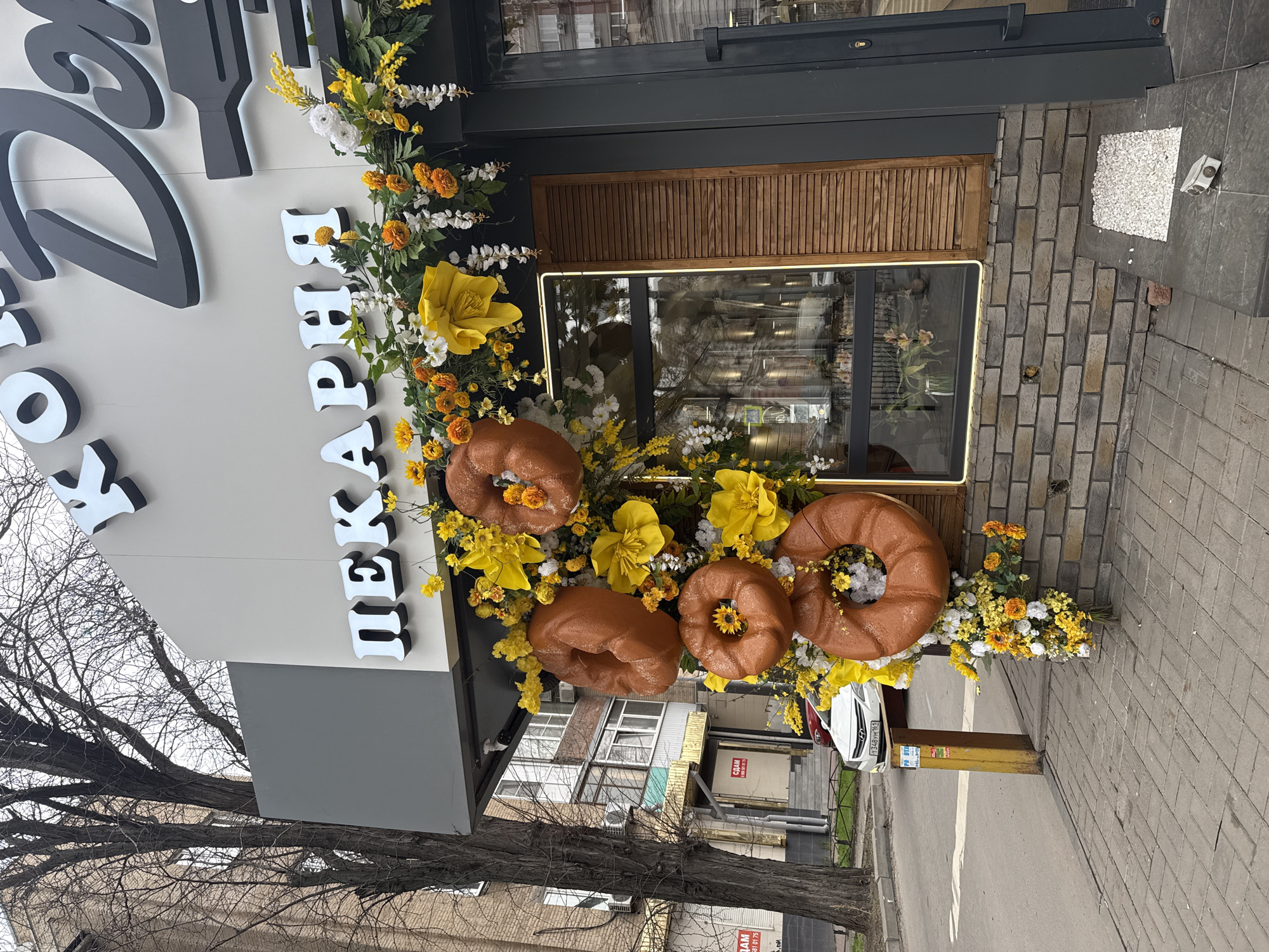 An eye-level, outdoor shot shows a building entrance decorated with flowers and large, donut-shaped sculptures in Rostov-on-Don, Russia.
The entrance is adorned with a cascade of yellow, white, and orange flowers, intermixed with four brown, glazed donut-shaped sculptures, two of which have sunflower centers. Above the floral arrangement, vertical white lettering spells out a name in Cyrillic script. To the left, a dark grey sign is visible.
The building exterior is composed of brick, and the doorway has a wooden shutter-style window and a dark grey frame. The window reflects the surroundings.
A tree with bare branches is partially visible in the foreground. A sidewalk runs along the side of the building, with a yellow pole and a car parked nearby. In the background, other buildings are seen.