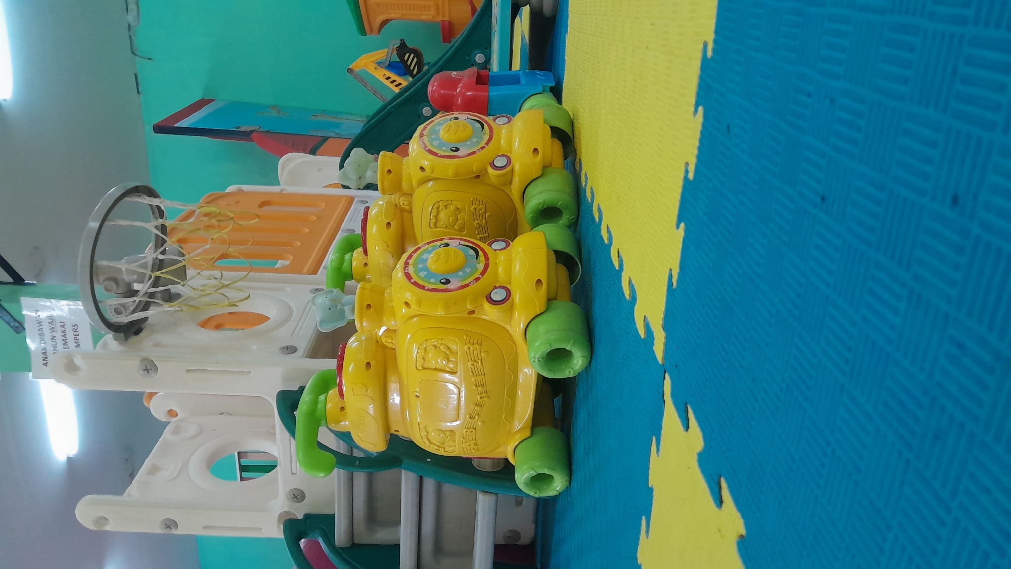 In Kasiman, Indonesia, this image captures a playful scene within an indoor children's play area. The foreground is dominated by a vibrant blue and yellow interlocking foam mat floor, designed for safety and comfort. Arranged in a row are three identical yellow toy trains, each with green wheels and an elephant figurine on top. These trains are facing forward, suggesting they are either waiting to be ridden or have just finished their use.
In the background, a multi-level plastic playground structure is visible, painted in white and orange. A basketball hoop with a yellow net is attached to the top of this structure, hinting at activities beyond just the trains. The walls of the play area are painted a bright teal color. A white sign with Indonesian text hangs from the ceiling, partially obscured by the playground equipment, indicating rules or information for the area. The lighting suggests an indoor environment, likely a daycare, kindergarten, or indoor playground facility. The overall impression is one of a cheerful and engaging space for young children.