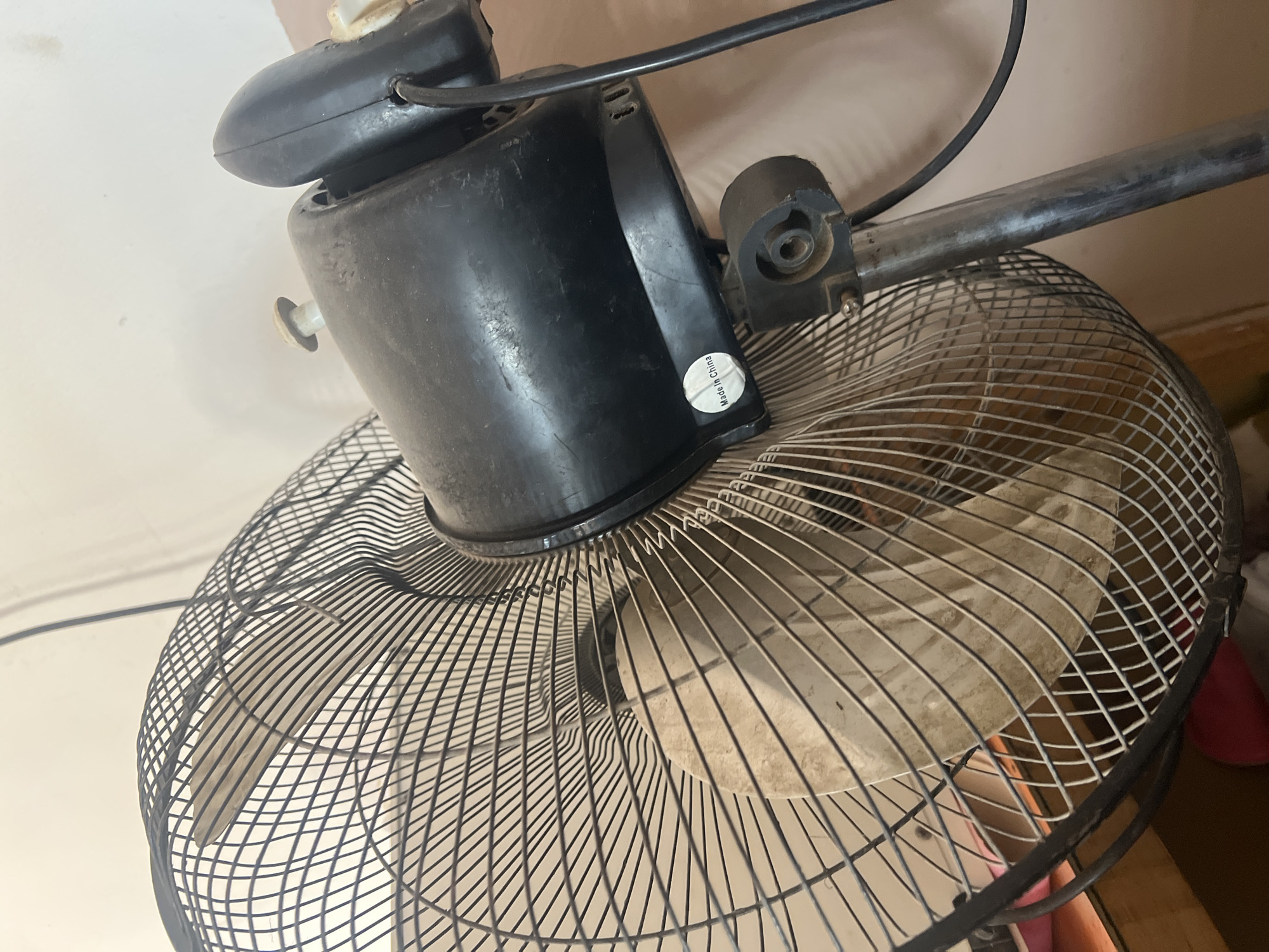 A close-up, angled shot shows a black, dusty oscillating fan with a metal grille and beige plastic blades. The fan is mounted on a metal pole that is partially visible. A small white circular sticker with "Made in China" printed on it is affixed to the black housing of the fan. The background is a plain, light-colored wall and a wooden surface, possibly a desk, with some blurred red objects underneath. The scene is indoors, and the lighting suggests it is daytime. The overall impression is of a functional but somewhat neglected appliance. The location of Jalingo, Nigeria, is not directly indicated by any visual cues in the image.