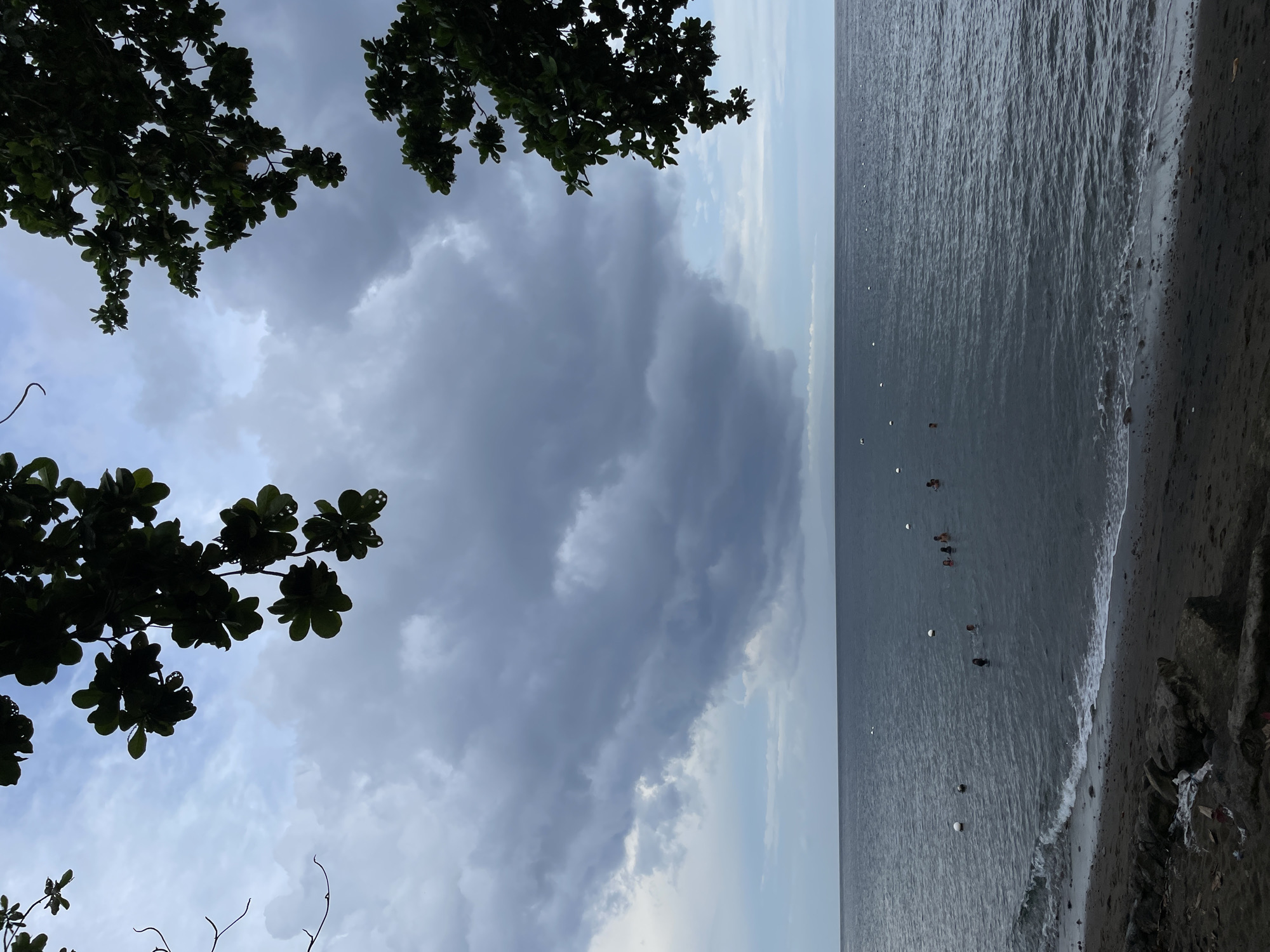 The image shows a dark, overcast sky over the ocean in Dauin, Philippines. Dense, dark clouds dominate the upper portion of the sky, with lighter clouds and a pale blue sky visible near the horizon. Silhouetted against the sky are branches and leaves of trees on either side of the frame. The sea is dark and choppy, with gentle waves breaking on a dark sand beach in the foreground. Several buoys are visible in the water, and a group of people, likely swimming, are scattered throughout the sea. The beach itself is littered with rocks and some debris.