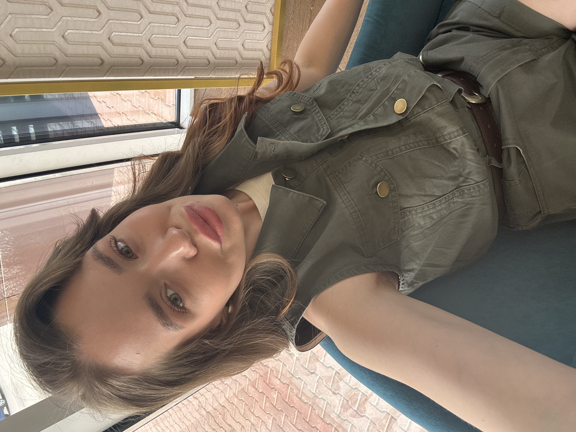 A young woman is seen in a close-up, low-angle shot. She has light skin, light brown hair, and green eyes. She is wearing an olive green outfit with a brown belt. The environment includes what appears to be an interior setting with a window visible in the background, suggesting a view from inside a building. There is patterned wallpaper or a textured window covering visible. The surface underneath her is teal. There is no explicit evidence to definitively confirm the location is Chaikovsky, Russia.