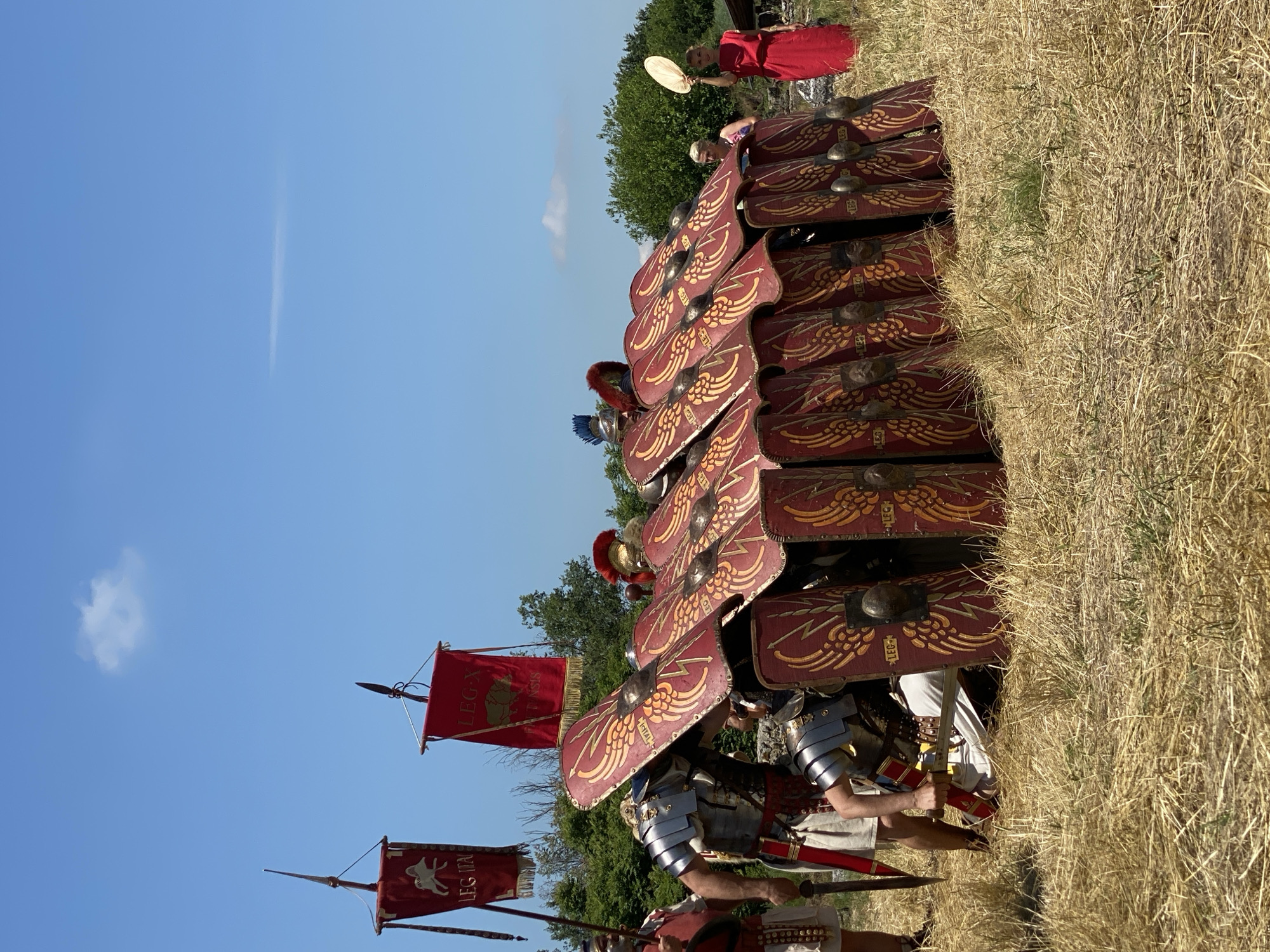 The image depicts a historical reenactment of Roman legionaries forming a *testudo* (tortoise formation) under a clear blue sky in Gigen, Bulgaria.
Approximately ten individuals dressed as Roman legionaries are arranged across the middle ground. Most are crouched or kneeling, interlocking large, rectangular, red and gold shields (scuta) to create a defensive formation. The shields feature a prominent golden-yellow winged design emanating from a central metallic boss, with the text "LEG" visible on multiple shields. Several legionaries wear segmented armor (lorica segmentata) and helmets, some with red or blue plumes. One legionary on the far left stands, clad in armor, holding a spear. Another legionary, lower and center-left, is seen adjusting something while still wearing armor and holding a gladius.
Two Roman military standards stand upright on spear-like poles in the upper-left quadrant. The left standard is red with a white animal emblem (possibly a ram or goat) and the text "LEG ITA". The right standard is also red, featuring a green map-like emblem and the text "LEG X FRETENSIS".
Two civilian women are observing the scene on the right side. The woman on the far right wears a simple red gown and stands facing left, holding a circular, light-colored object (possibly a fan) aloft in her right hand. Another woman is partially visible behind the shields, to the left of the woman in red.
The scene takes place outdoors in a dry, straw-colored grassy field. A dense line of green trees and bushes forms the background horizon. The sky above is a uniform clear blue, with a single small, white cumulus cloud visible in the upper-center-left. A faint, thin, wispy cloud streak is visible further to the right. A dark, wooden structure or ruin is partially visible on the far right, behind the woman in red.