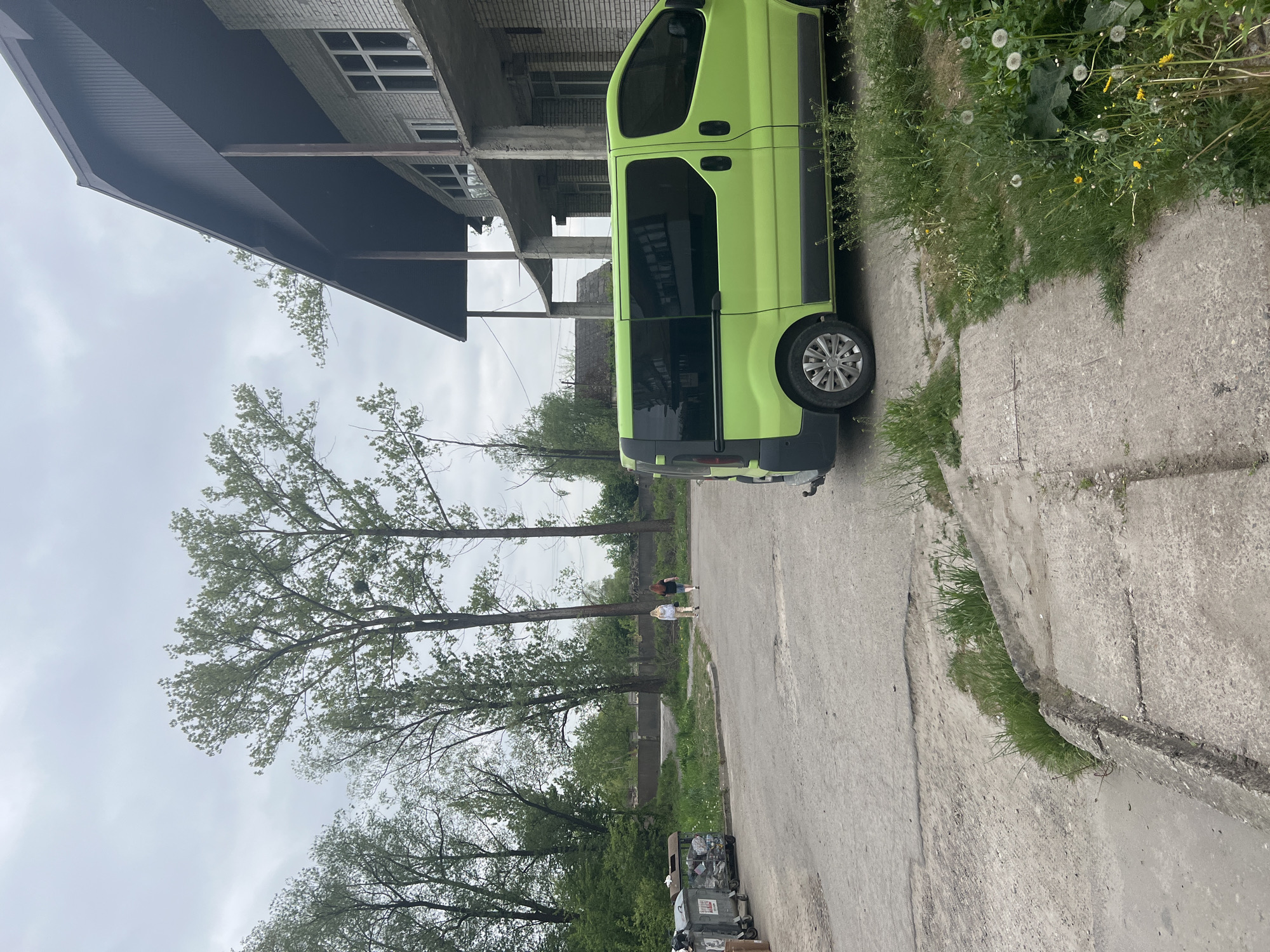 The image shows a bright green van parked on a concrete street in what appears to be an urban or suburban area of Cherliany, Ukraine. The van is parked next to a curb with grassy areas on the other side, interspersed with dandelion-like plants. A building with multiple stories and unfinished construction is visible behind the van. Trees are in the background, and two individuals are walking in the distance. Several trash bins are visible along the street. The sky is overcast.