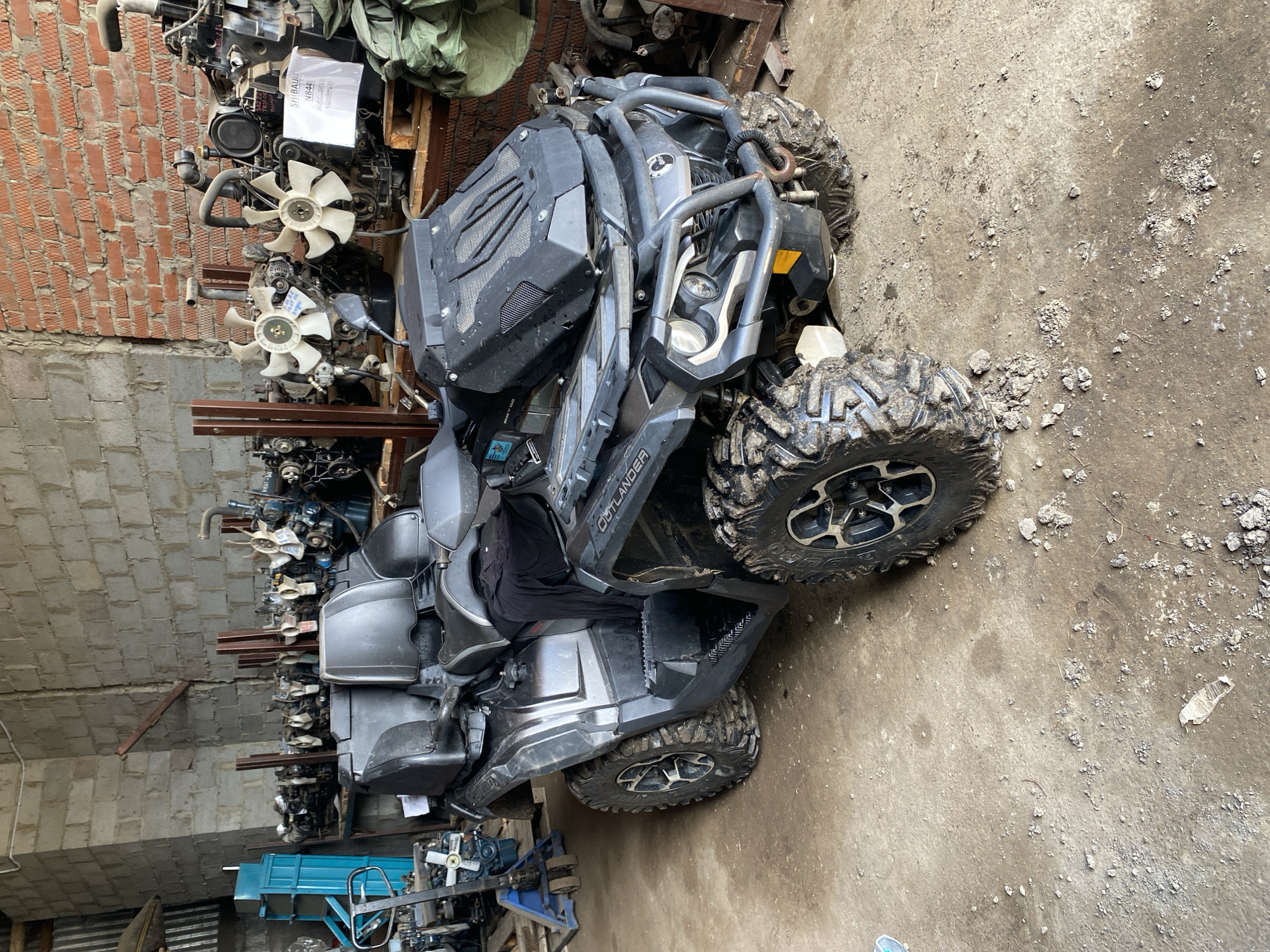 A black and gray Outlander ATV is parked indoors on a concrete floor. The surroundings suggest a workshop or storage area. To the left, shelves are stacked with various mechanical parts, including engines and fans. A brick wall is partially visible in the upper-left corner. The floor is covered with dirt and debris. The image does not contain explicit location information beyond the possible city mentioned.