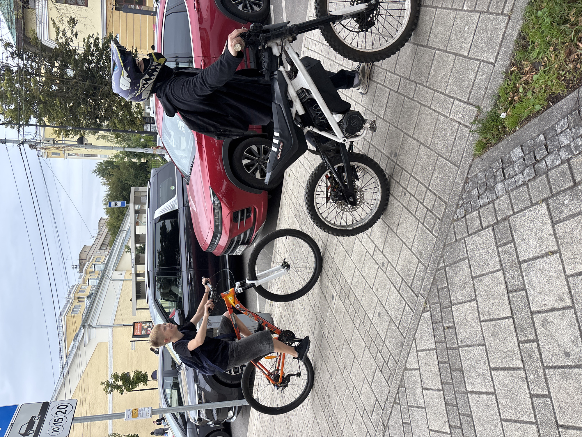 In Zvenigorod, Russia, two individuals are pictured on a sidewalk adjacent to a street with parked cars and buildings in the background. The foreground features a young boy on an orange bicycle, wearing a black t-shirt and shorts, his expression suggesting enjoyment. To his right stands a person wearing a black hoodie, a distinctive helmet with a sharp design, and dark pants, holding a white electric bicycle. The electric bicycle has a robust frame and off-road tires, indicating a more rugged design. The pavement is a mosaic of grey and darker grey rectangular tiles, with a strip of cobblestones at the edge. The environment suggests a mixed urban and possibly semi-residential area, with a cloudy sky overhead. Signage is visible in the upper left corner, with the numbers "10 15 20" and a car icon, possibly related to parking or traffic restrictions. Further background elements include trees, more parked vehicles, and buildings with light-colored facades.