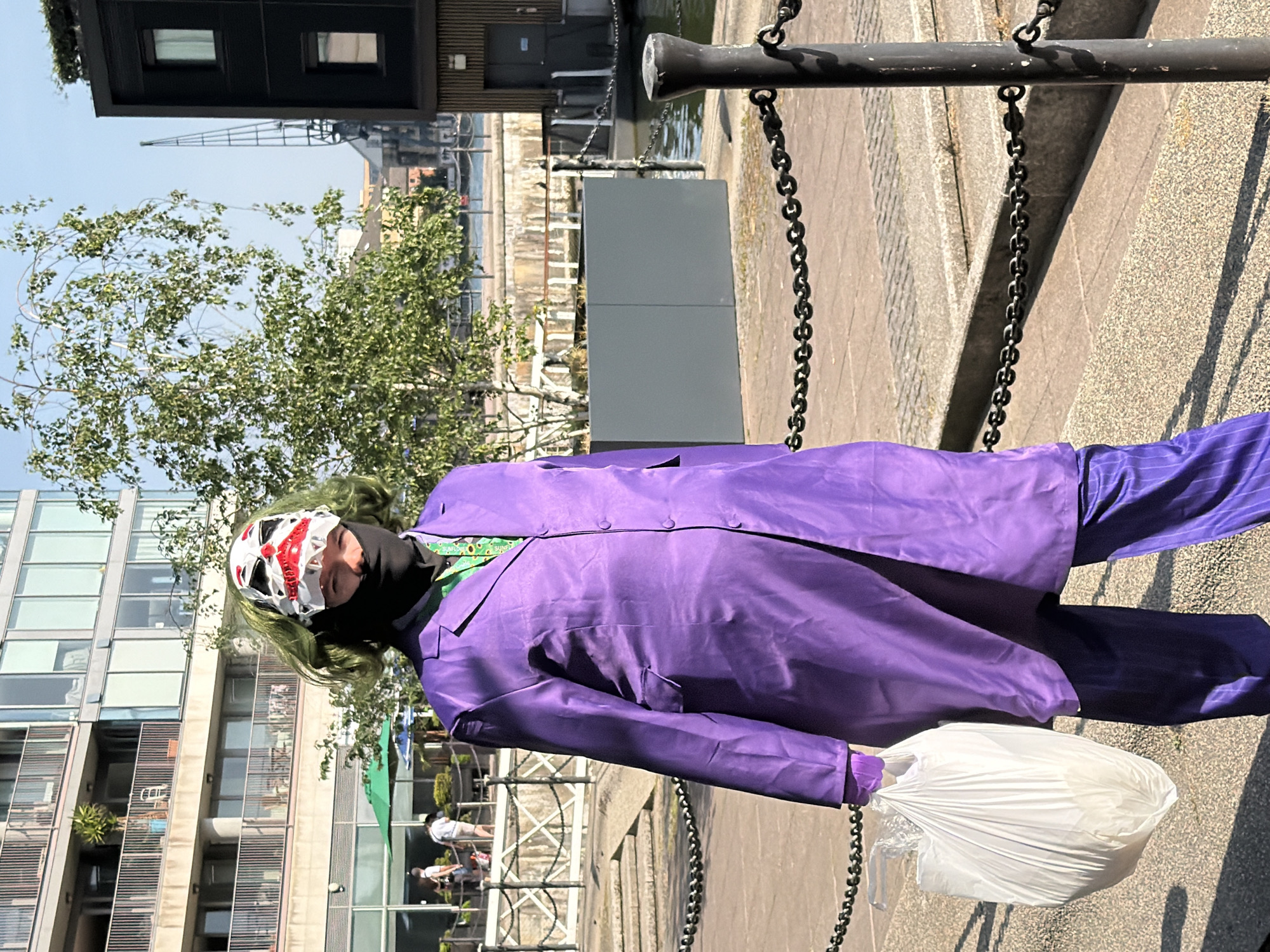 A person dressed as the Joker is standing outdoors in London, United Kingdom. The person is wearing a purple suit, green hair, and a mask. They are holding a white plastic bag. The background includes modern buildings, a tree, a harbor with a crane, and a section of a metal chain link fence. The time of day appears to be daytime.