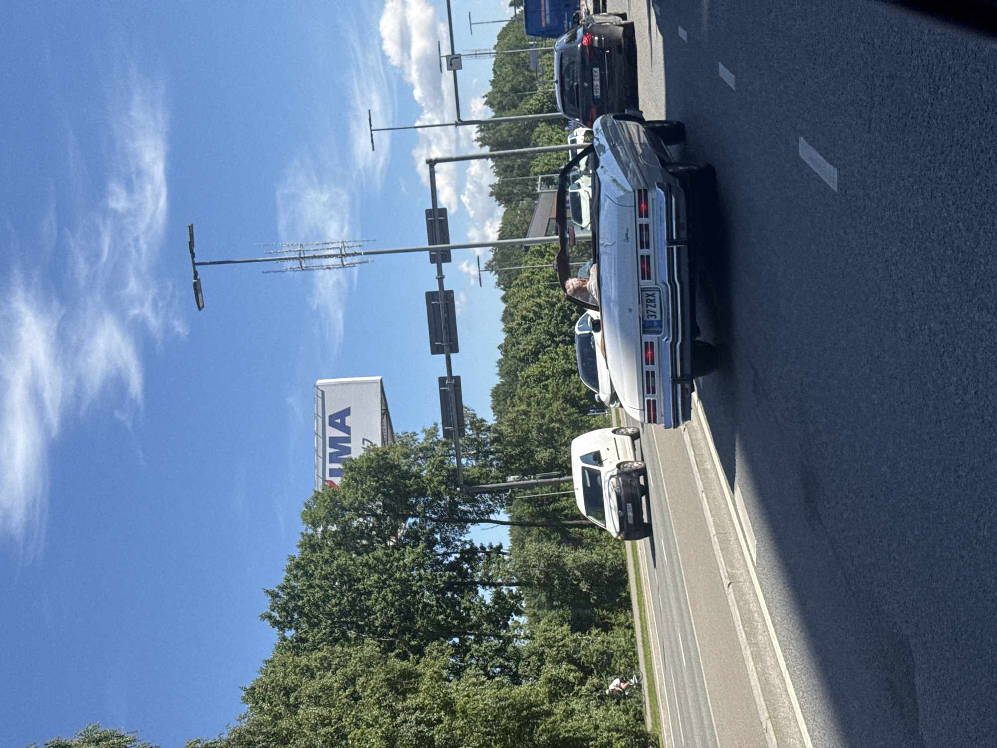 In Tallinn, Estonia, on a sunny day, traffic moves along a multi-lane road bordered by dense green trees on the left. The foreground shows the asphalt road surface with lane markings and a shadow cast from an unseen object. In the middle ground, several vehicles are visible. A white van is in the left lane, followed by a black sedan in the right lane. In the center of the road, a classic white convertible is driving, with a person with blonde hair visible behind the wheel. This car has a license plate that reads "37 ZRX". A blue truck is partially visible in the far right lane. In the background, a large billboard with the word "MA" is mounted on a tall structure, and traffic lights and streetlights are present, indicating an urban or semi-urban environment. A cyclist is also visible on the left side of the road.
