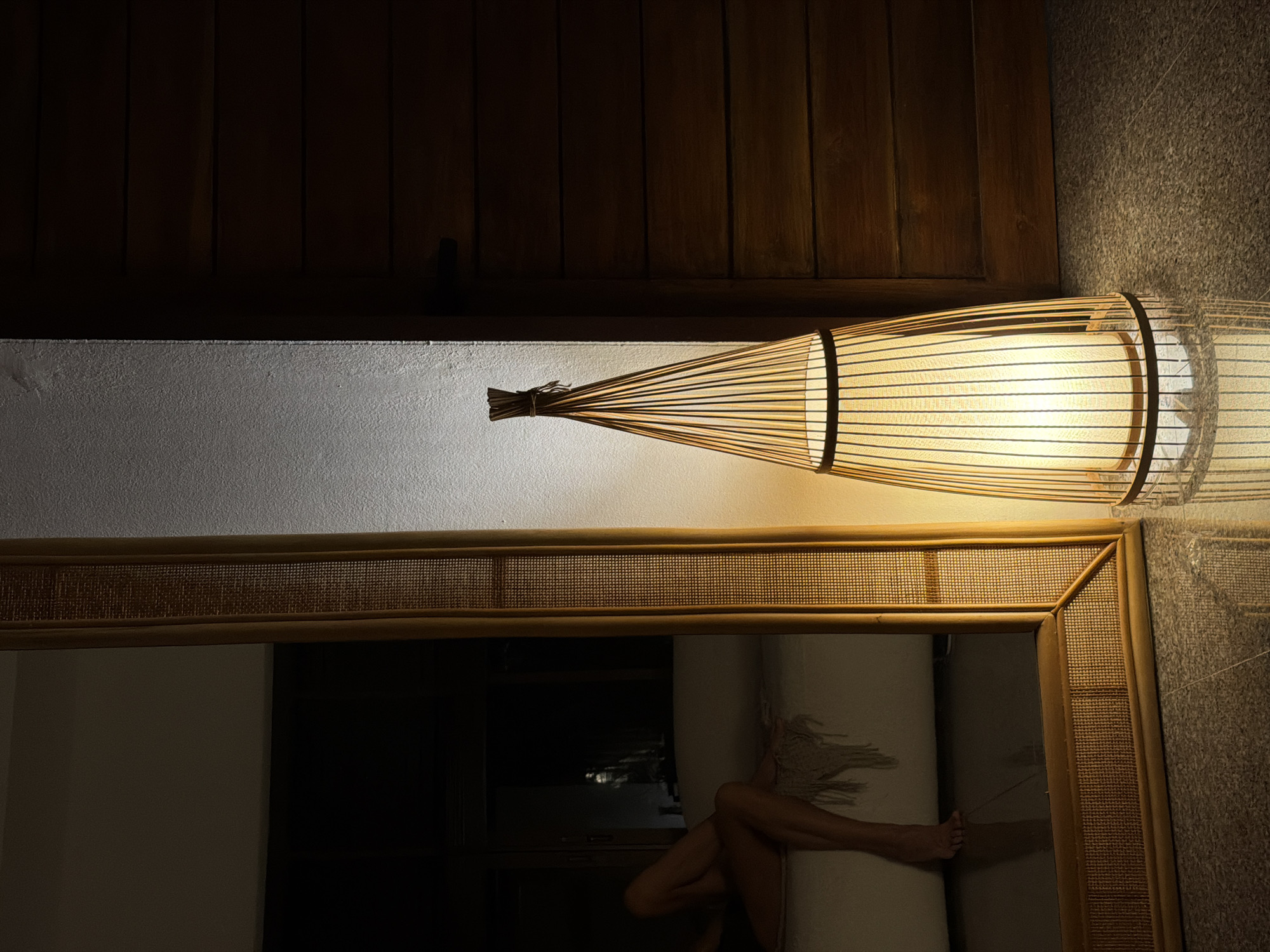 In a dimly lit room in Baan Bo Phut, Thailand, a tall, conical floor lamp with a woven bamboo shade is illuminated, casting a warm glow on the textured wall and the dark wooden door beside it. The lamp's base is surrounded by its reflection on the glossy tiled floor. To the left, a large, rectangular mirror with a rattan frame reflects a partial view of the room. In the reflection, a person's legs and feet are visible, resting on a white sofa with a fringed throw. The overall atmosphere is serene and intimate, with the play of light and shadow creating depth and ambiance.
